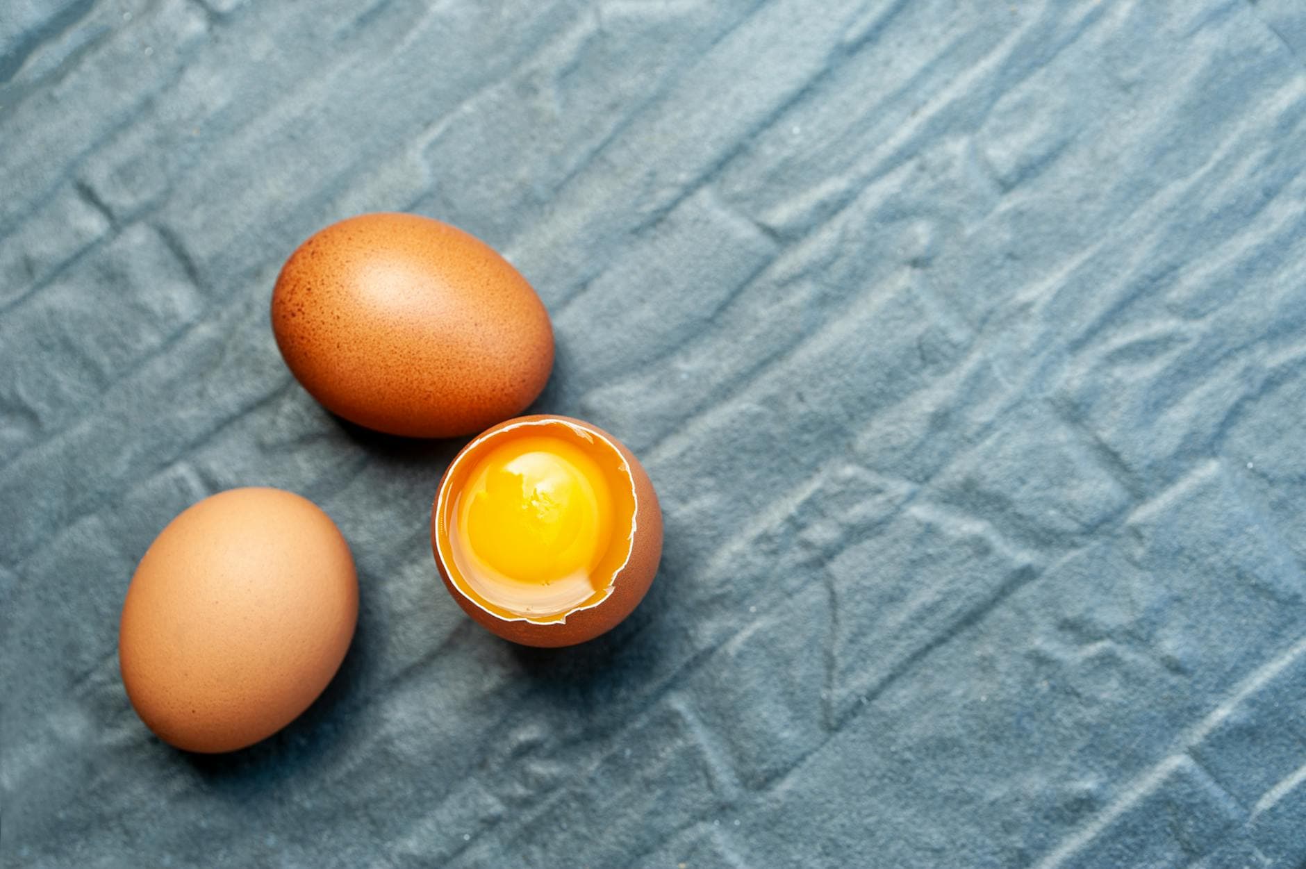 Close-up of three brown eggs on textured blue fabric; one egg is cracked open revealing the yolk. - healthy breakfast eggs