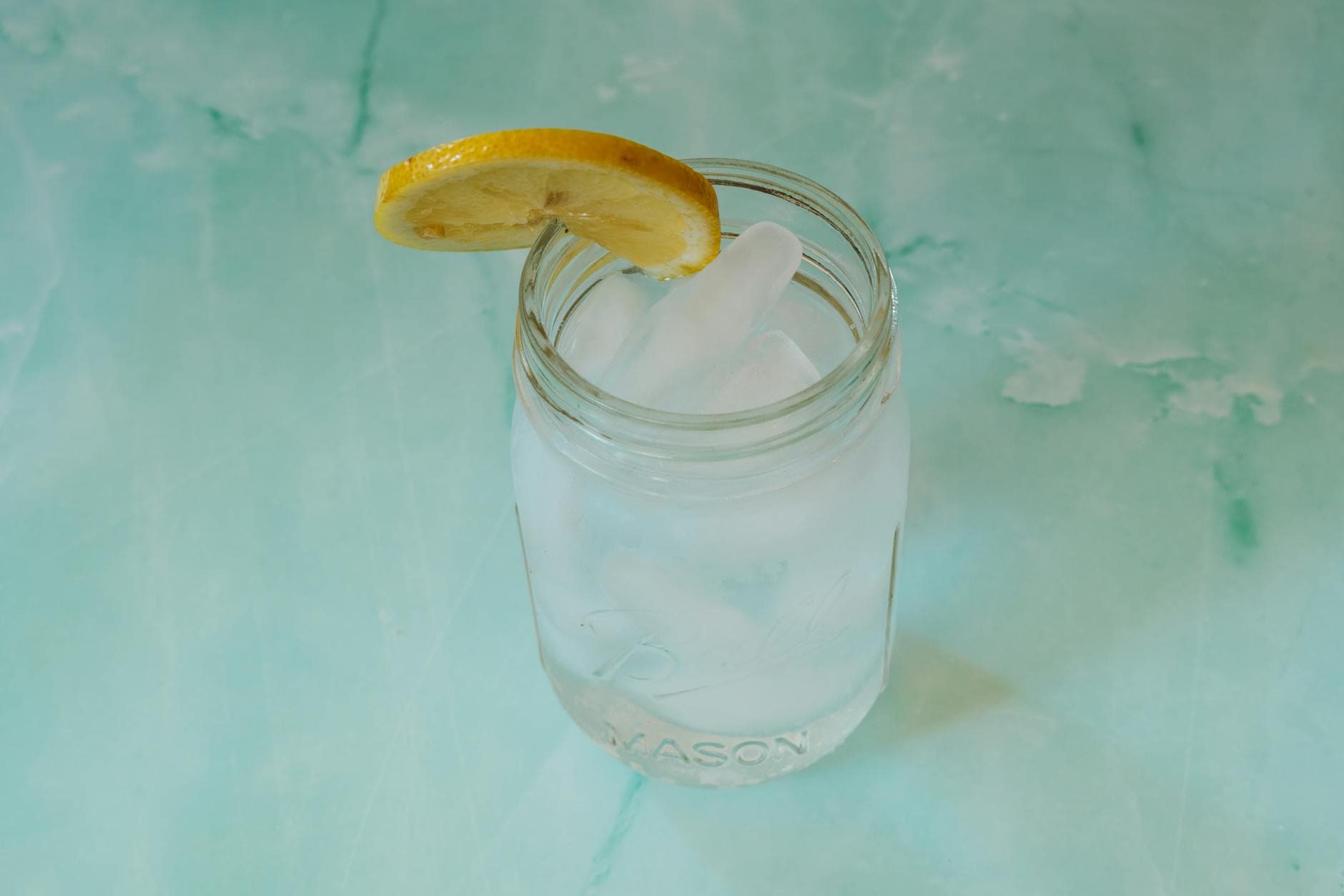 Close-up of a refreshing iced water in a mason jar with a lemon slice on a turquoise background, ideal for summer themes. - best detox water