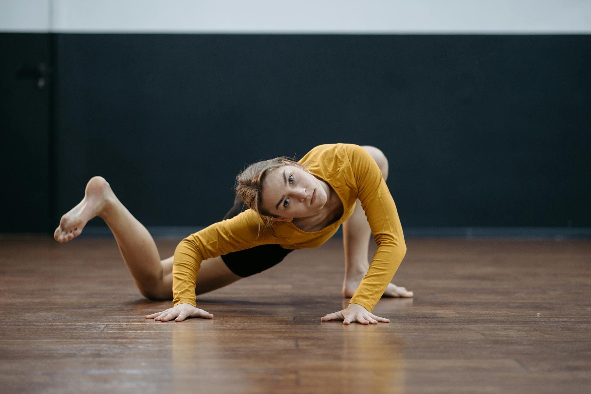 A woman in a yellow shirt performing a stretching exercise on a wooden floor indoors. - flexibility exercises list