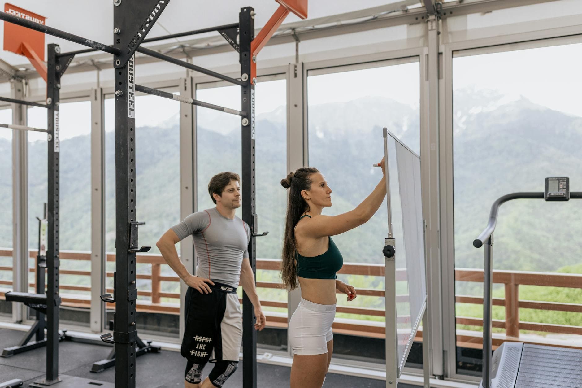 A gym coach instructs on whiteboard beside fitness equipment with mountain view. - gym workout plan