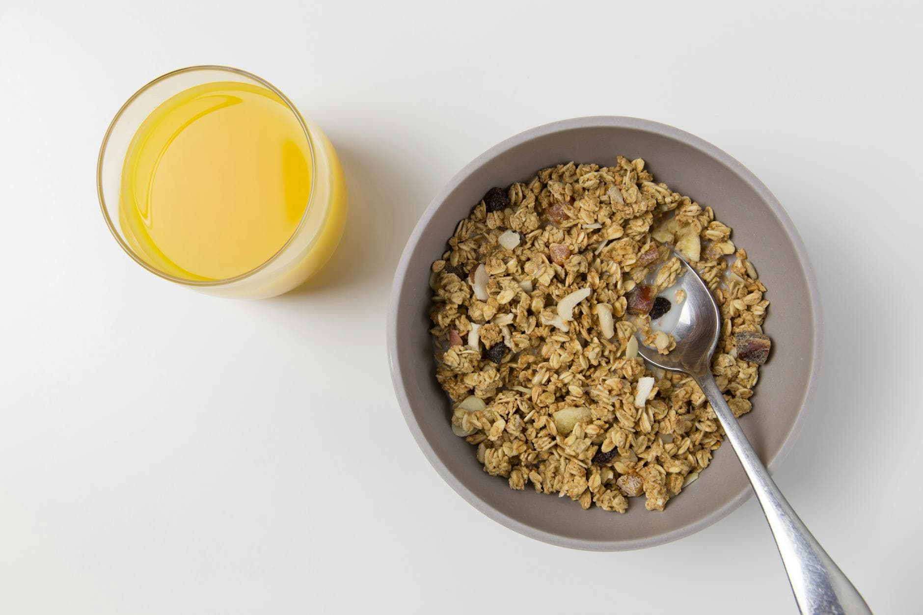 A top view of granola in a bowl with a spoon and a glass of orange juice on a white background. - healthy breakfast ideas
