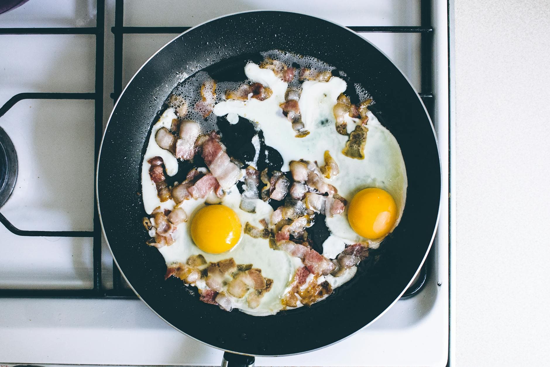 High-angle view of fried eggs and bacon sizzling in a skillet on a gas stove, perfect breakfast scene. - high protein breakfast ideas