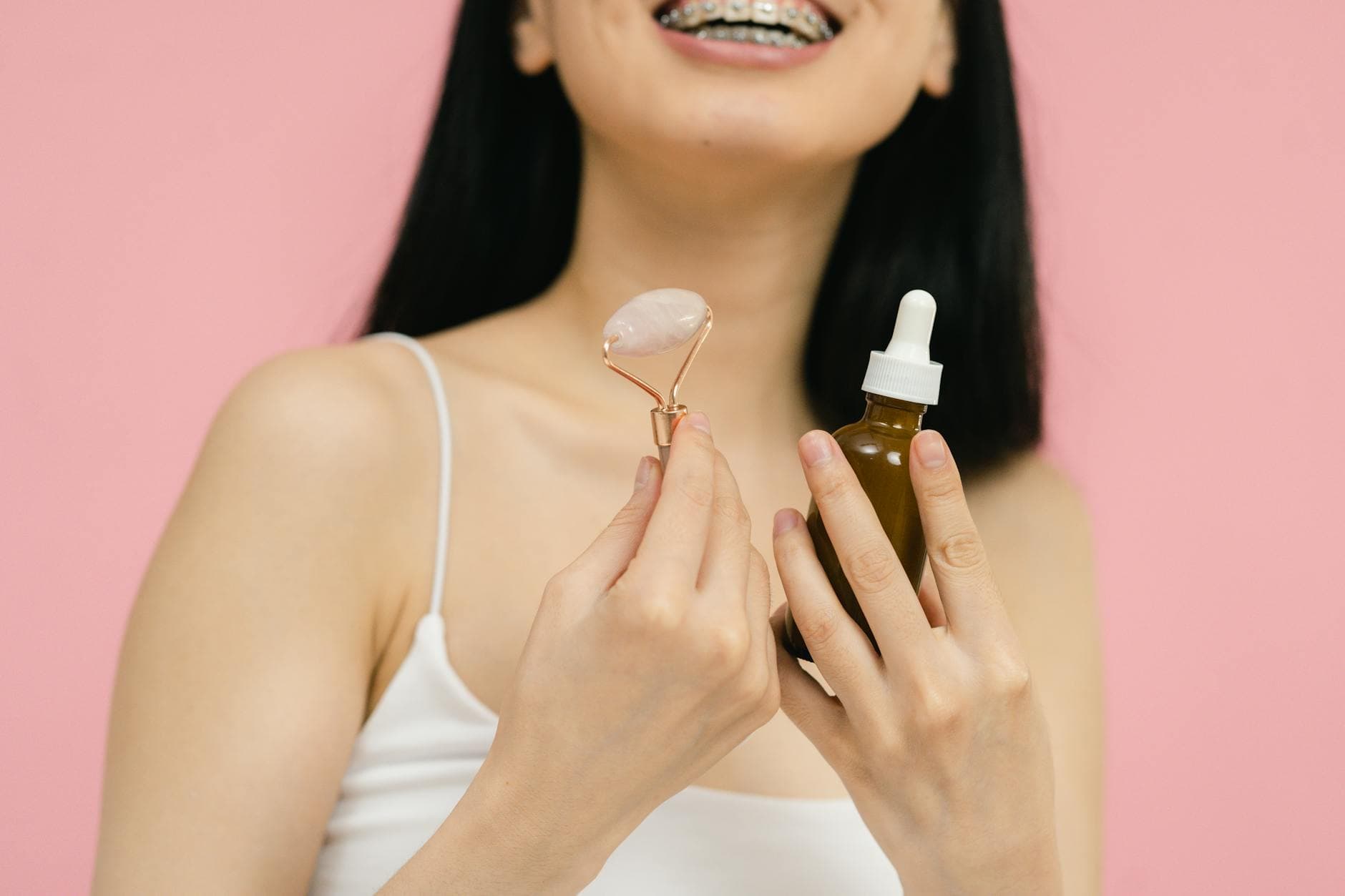 Smiling woman with braces using a facial roller and serum for skincare on a pink background. - radiant skin foods