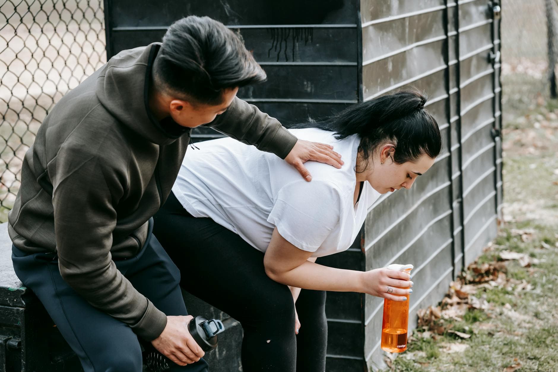 Sportsman clapping shoulder of plump tired female trainee sitting near net fence with bottle of water after training in autumn park - break weight loss plateau