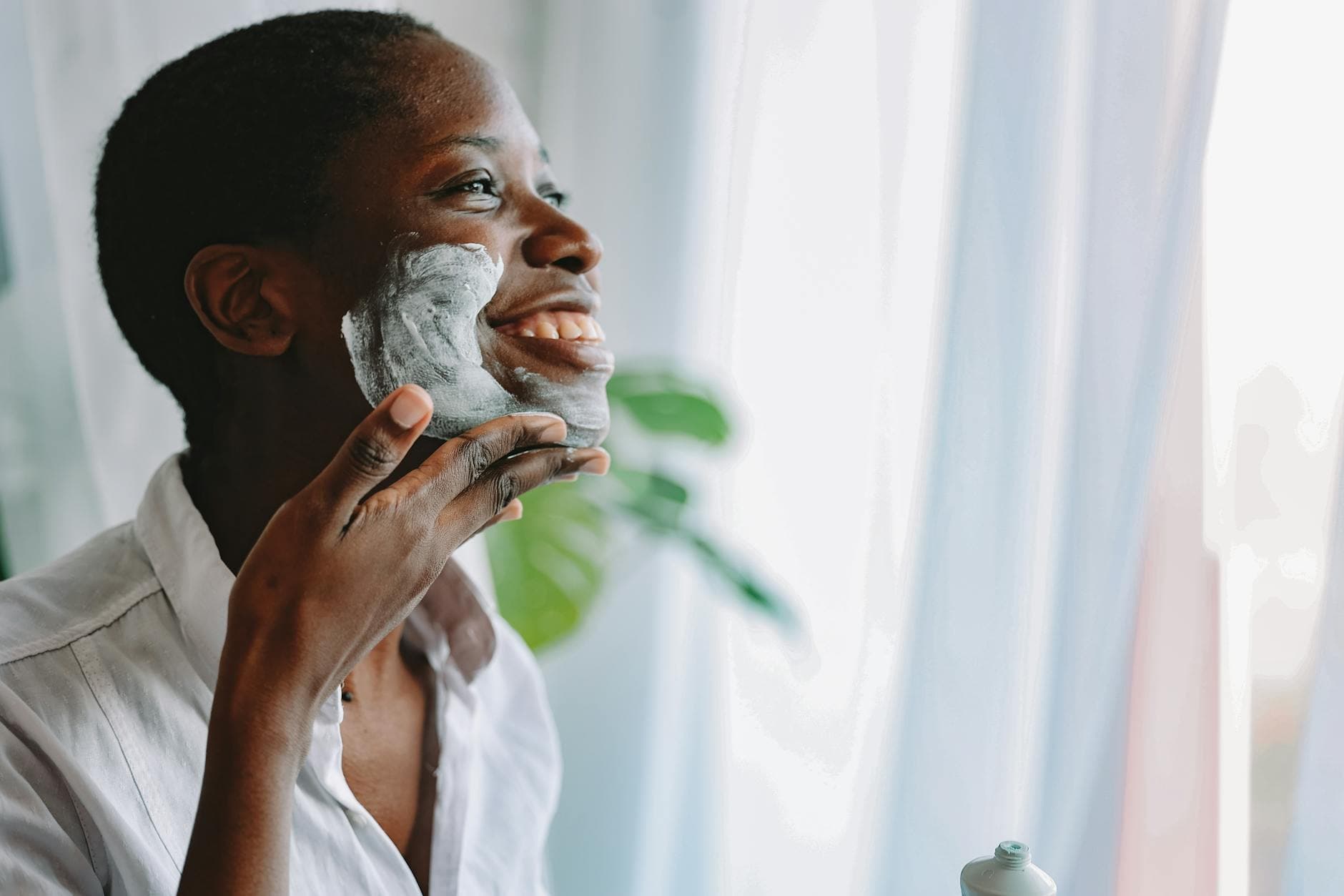 An African woman smiles while applying skincare cream to her face indoors. - daily skincare routine