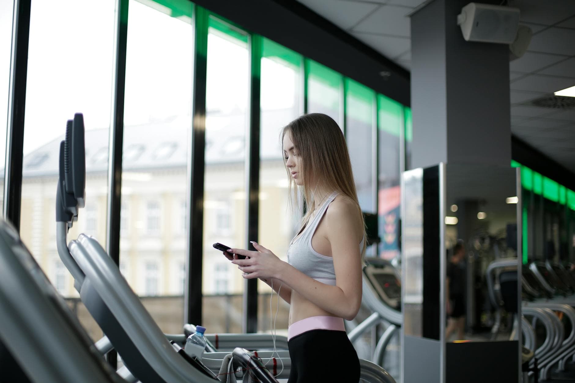 Side view of slim young female athlete in sportswear standing with smartphone and training on treadmill in spacious modern fitness center - interval training cardio