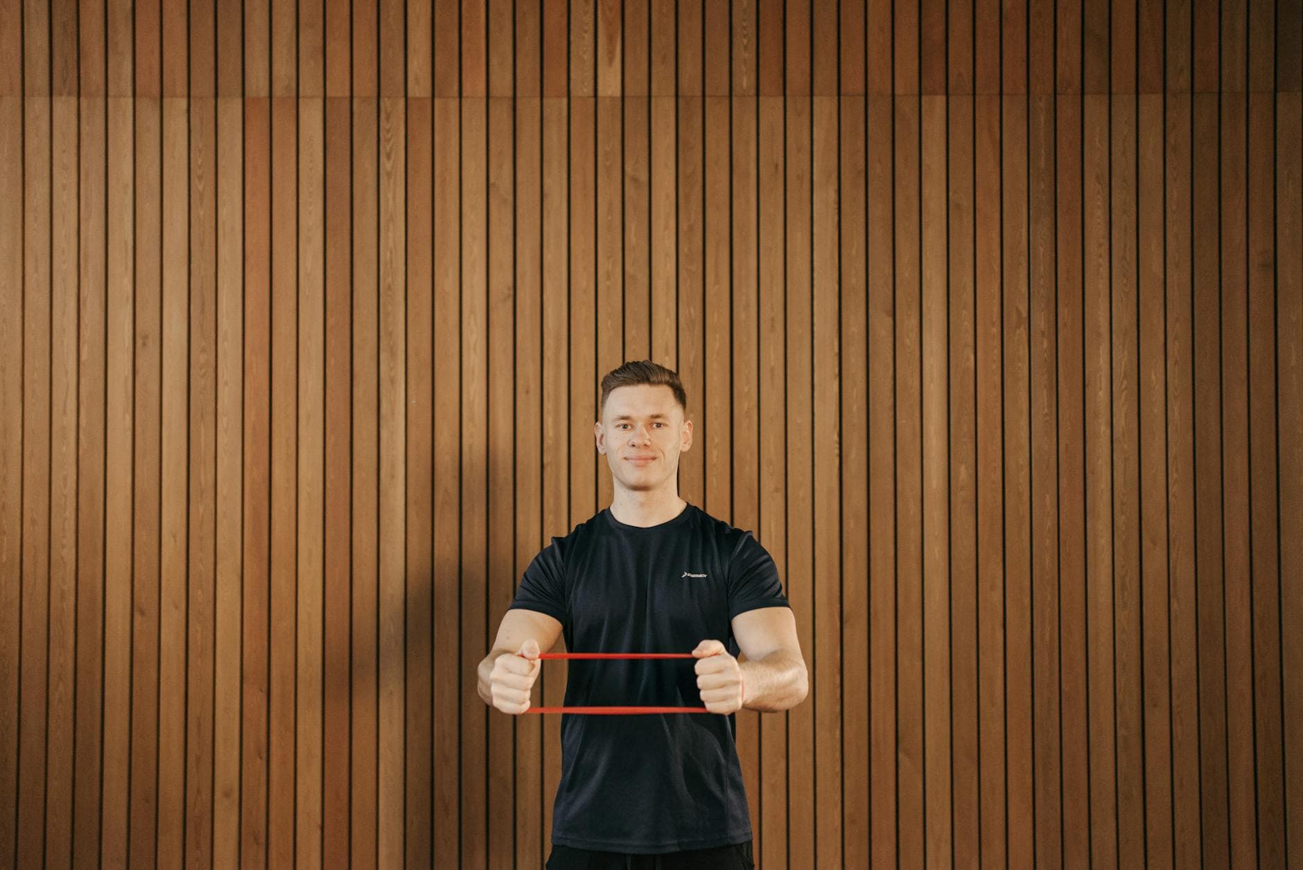 Man in a black shirt using a resistance band for exercise against a wooden wall indoors. - posture exercises reddit