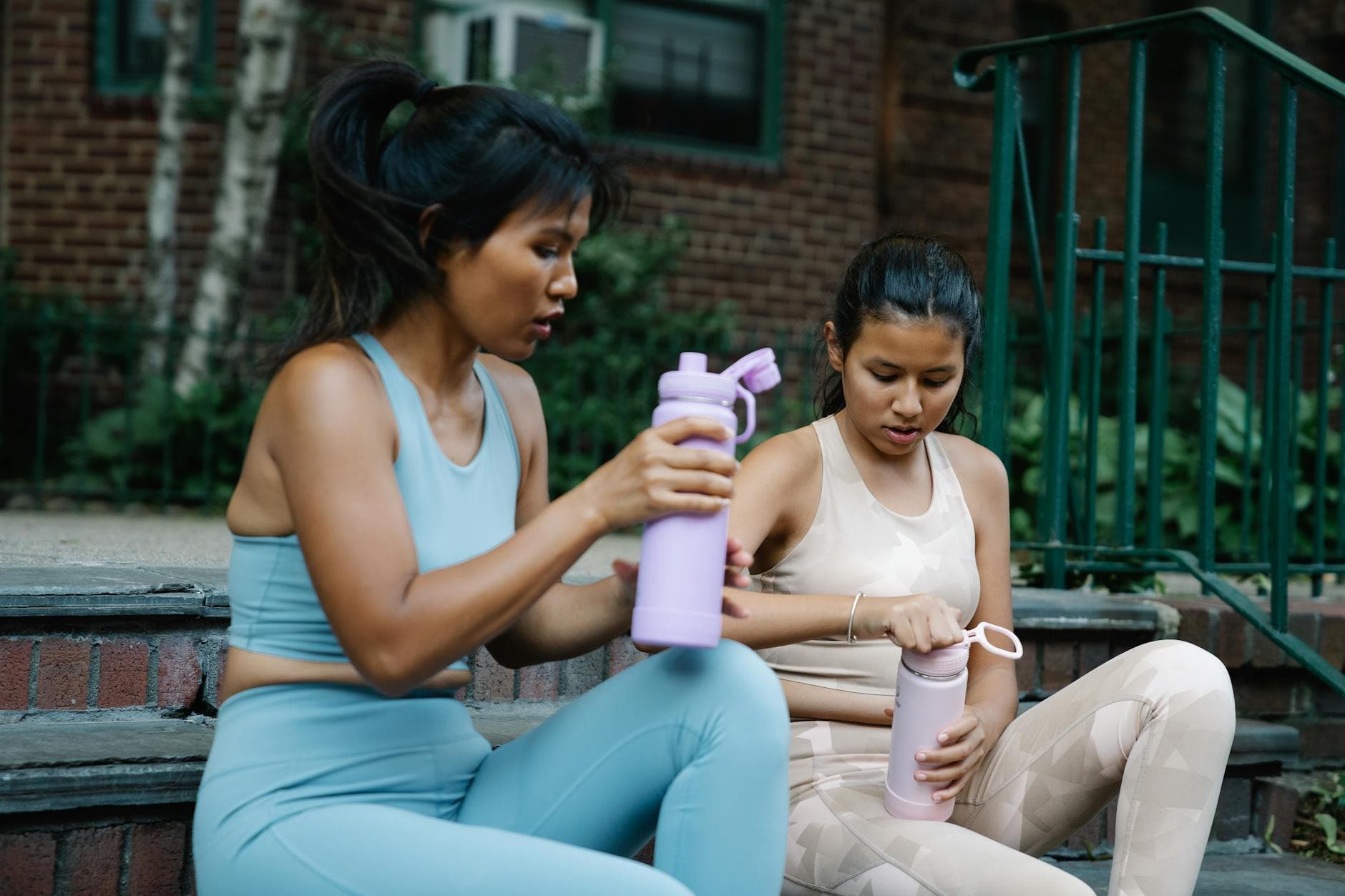 Two women in active wear resting on stairs, holding tumblers. Outdoors on a city street. - spring workout hydration