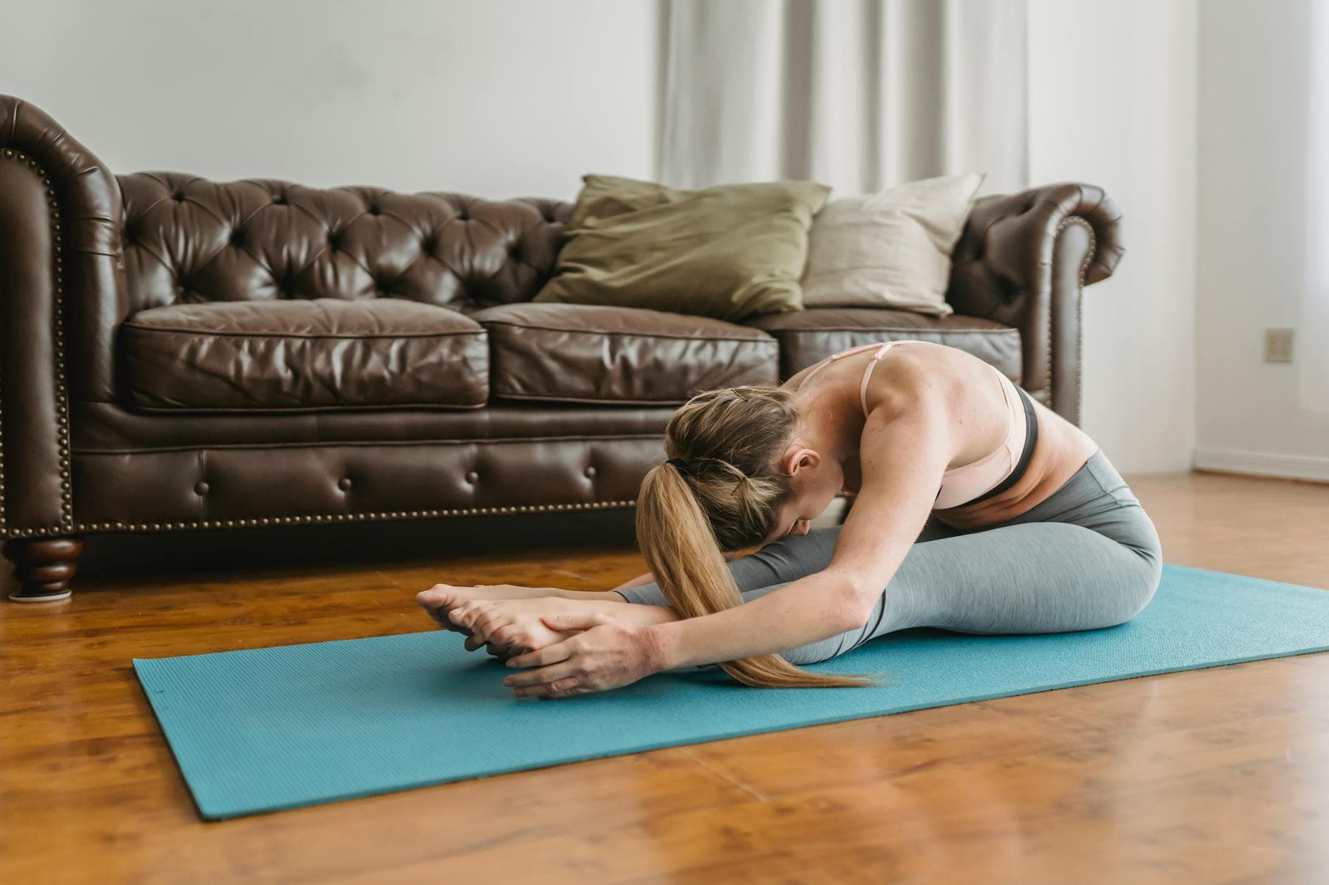 Side view of slim young lady with long blond hair in sportswear recreating in Seated Forward Bend asana after yoga session in living room - improve posture