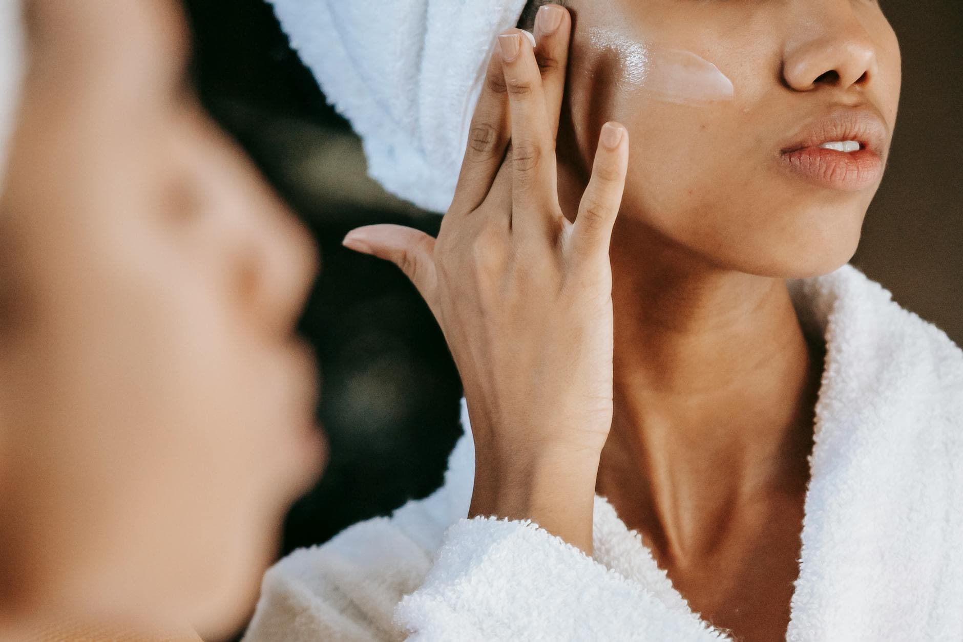 Crop unrecognizable young ethnic female in terry robe applying moisturizing cream on cheek while reflecting in mirror - nourish skin from within