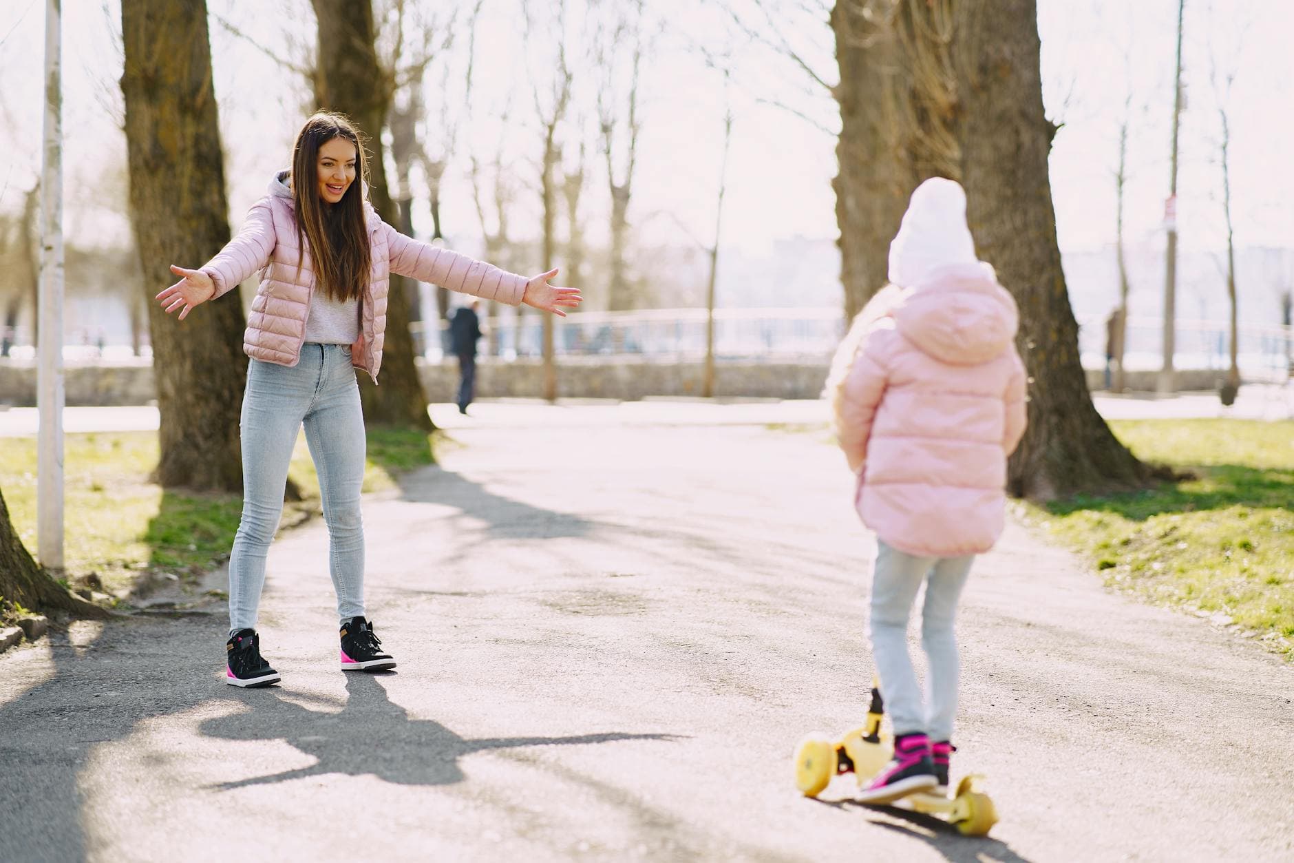 Full body little girl riding kick scooter on alley in park while mother standing with hands apart for support in sunny day - outdoor spring activities