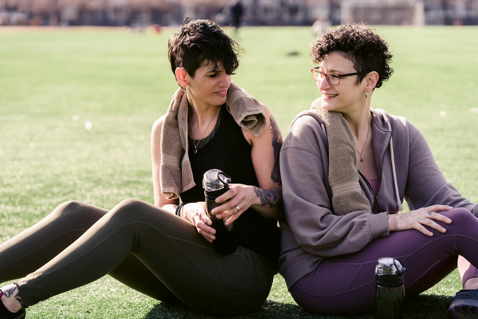 Cheerful female friends in activewear with bottles of water looking over shoulder at each other while sitting back to back on grass during training - outdoor workout hydration