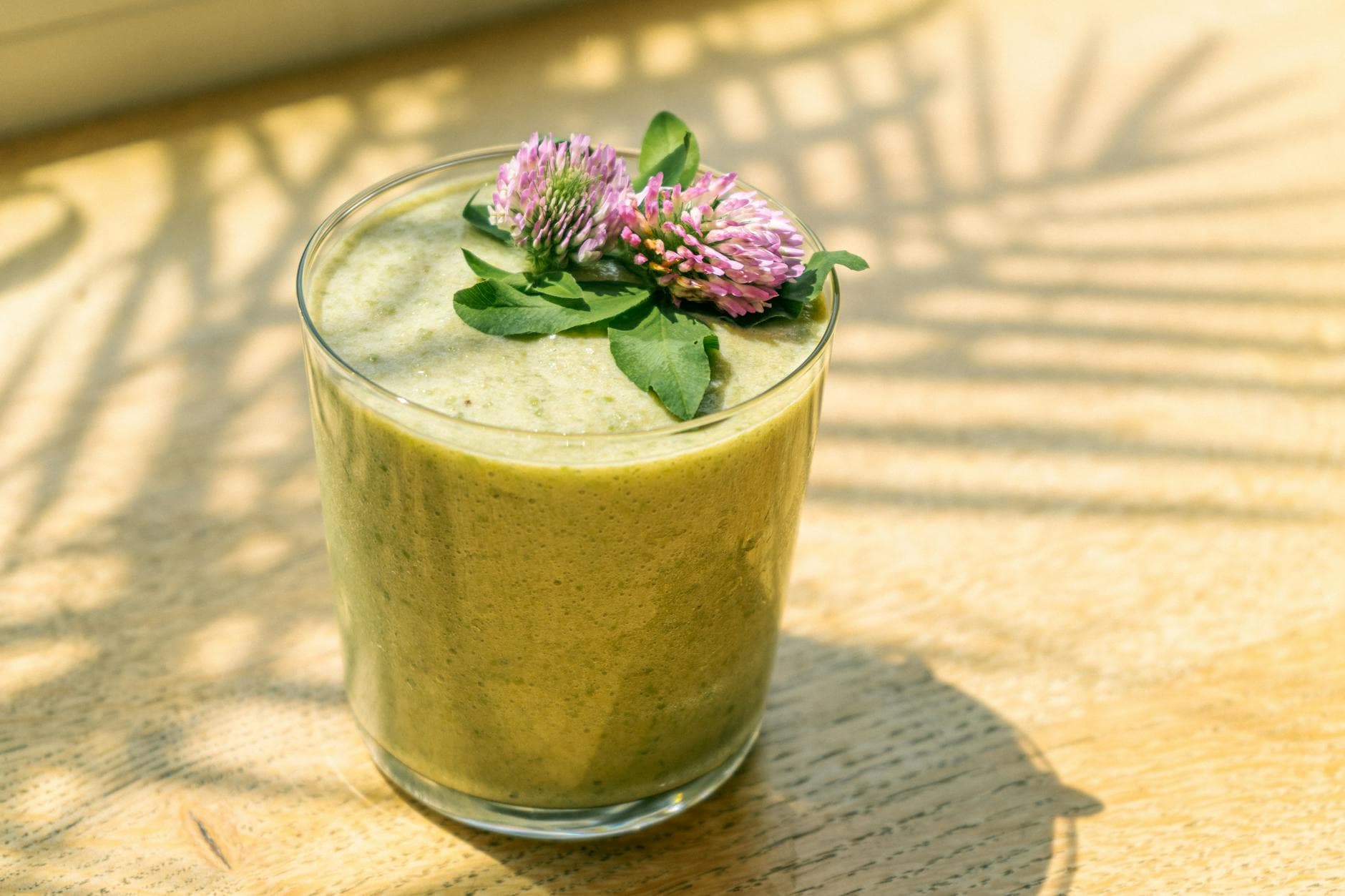 Close-up of a nutritious green smoothie topped with fresh purple clover flowers in sunlight. - spring detox smoothie