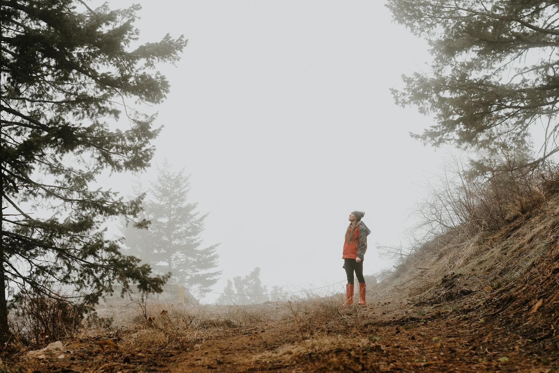 Full body of young female in rubber boots jacket and hat standing in hilly valley with evergreen trees in hazy day - spring hiking essentials