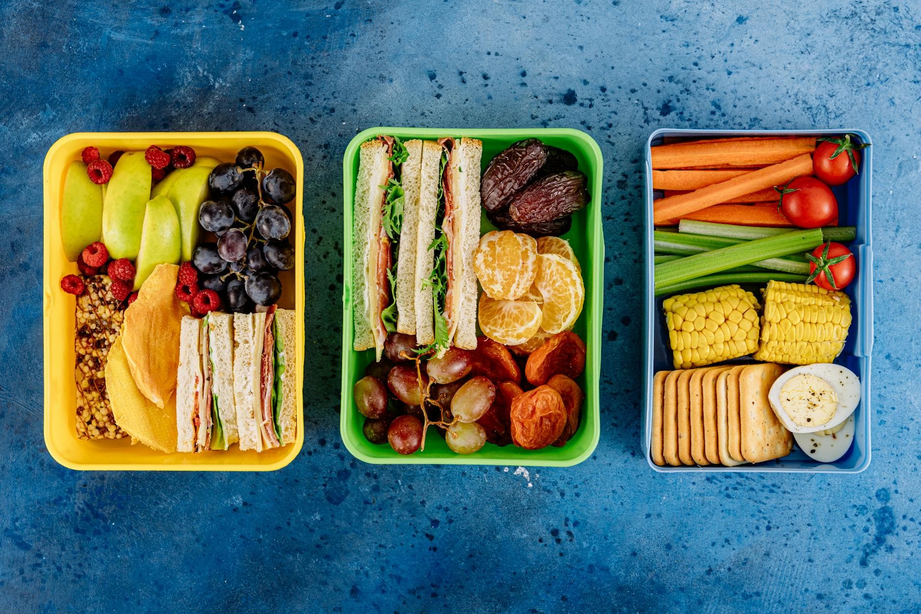 Three vibrant lunch boxes showcasing fruits, sandwiches, and vegetables on a blue textured background. - healthy work lunch ideas