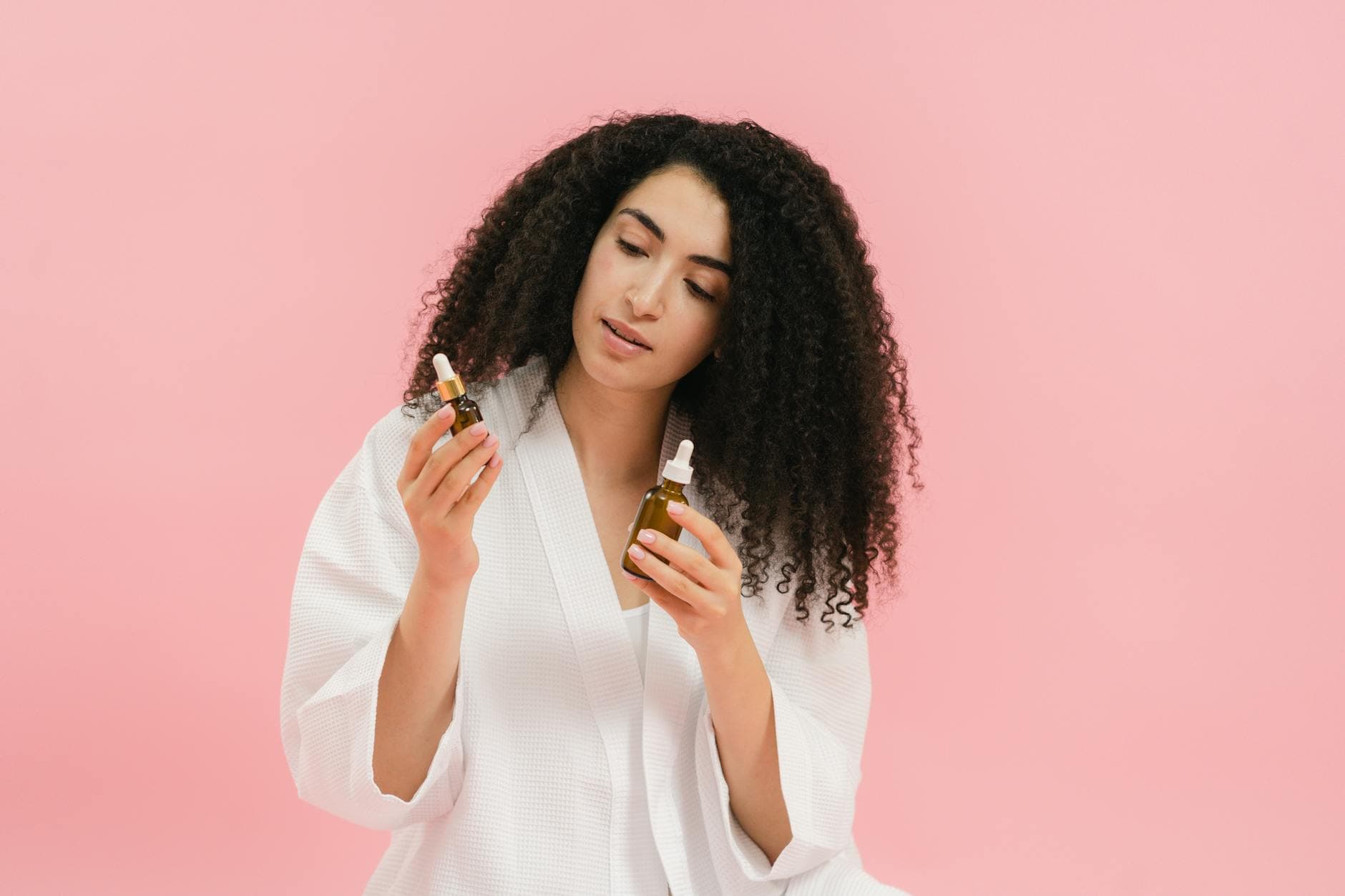 A woman with curly hair in a white robe examines two cosmetic oil bottles against a pink background. - just natural skincare reviews