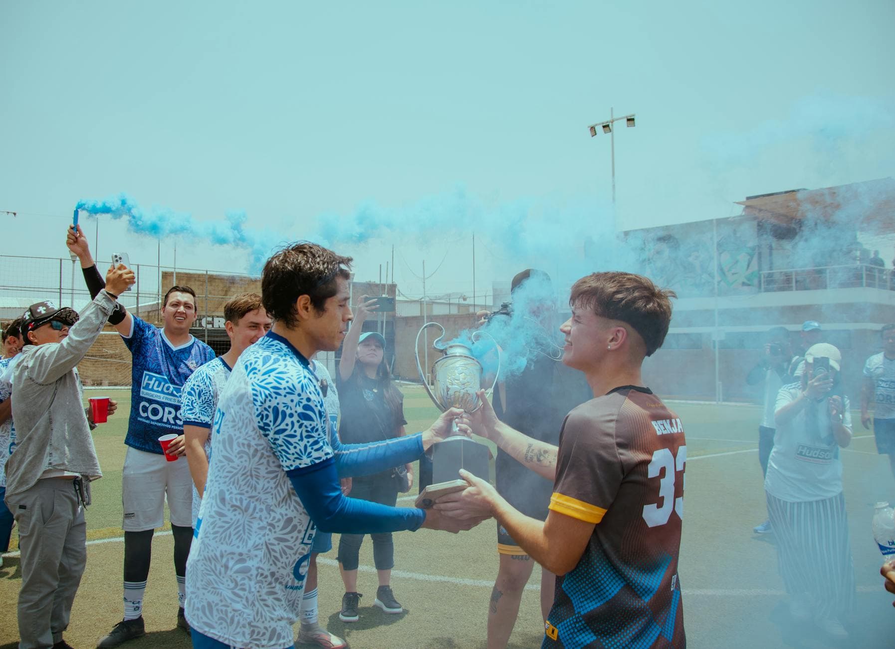 Soccer team celebrating victory with trophy, surrounded by blue smoke on outdoor field. - non-scale victories