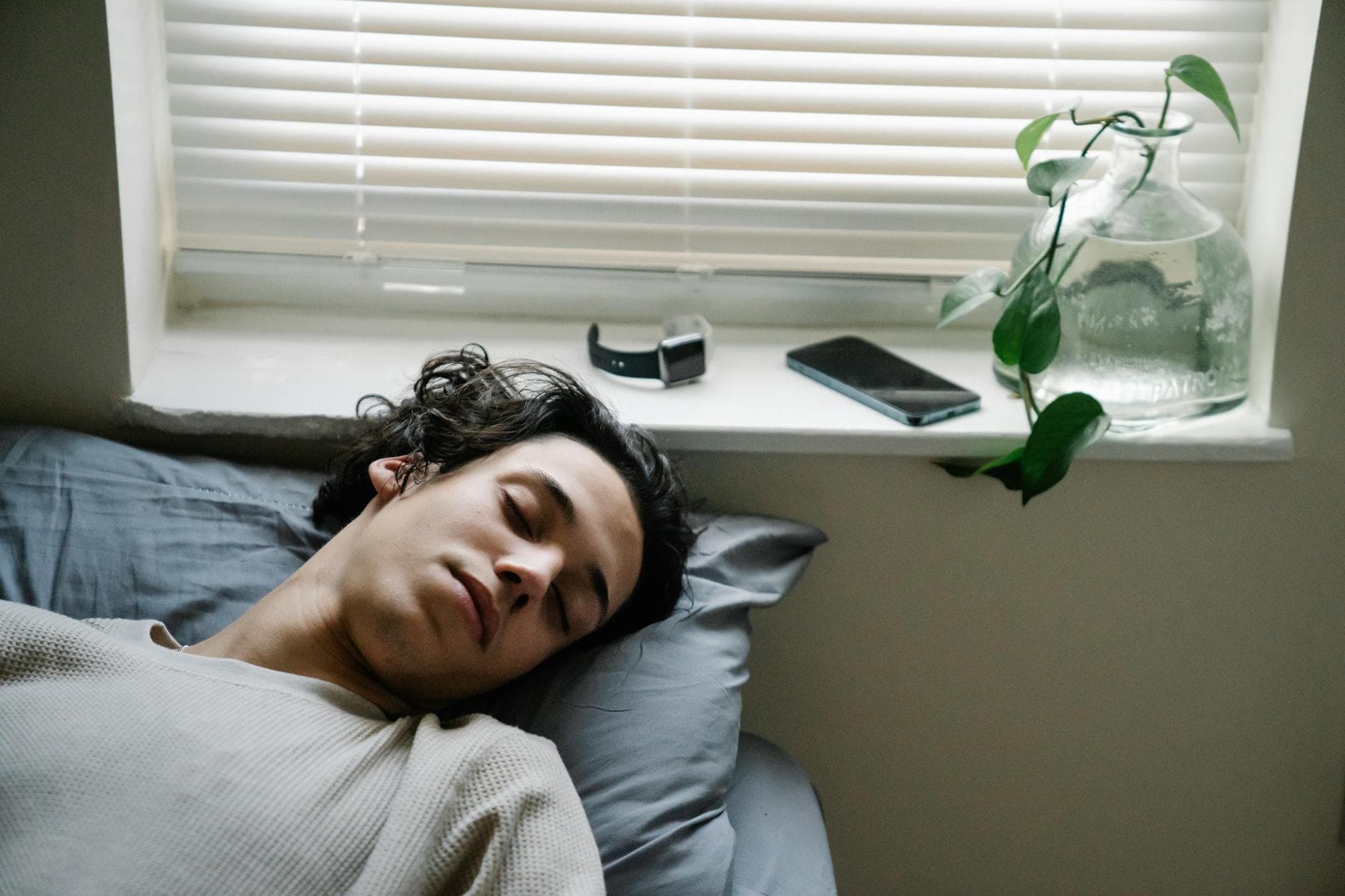 High angle of young ethnic male sleeping in bed with smartphone and smart watch placed near head on windowsill - sleep cycle reset