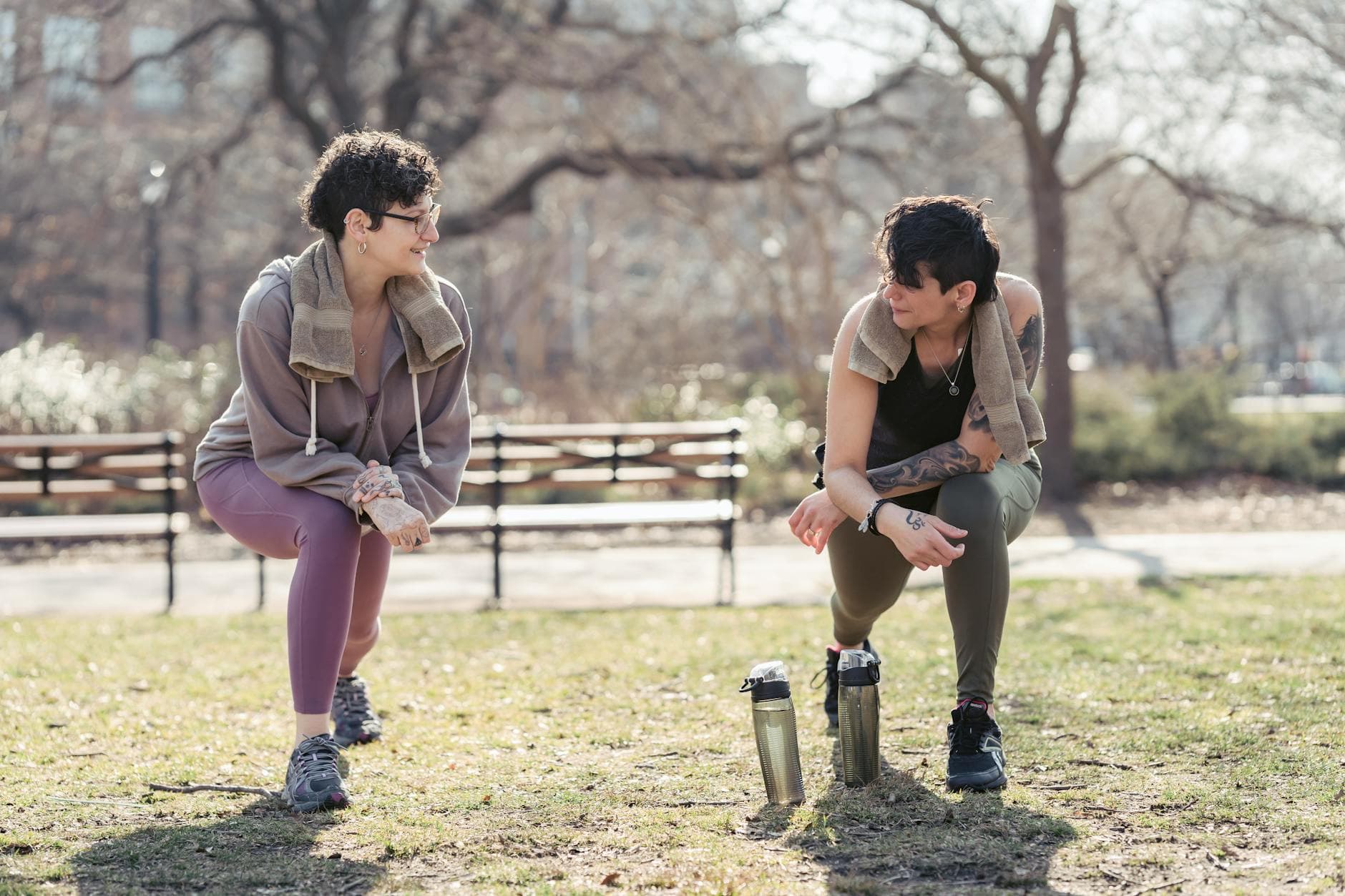 Full body of glad sportswomen in sneakers doing exercises in sunny park with leafless trees - spring wellness trends