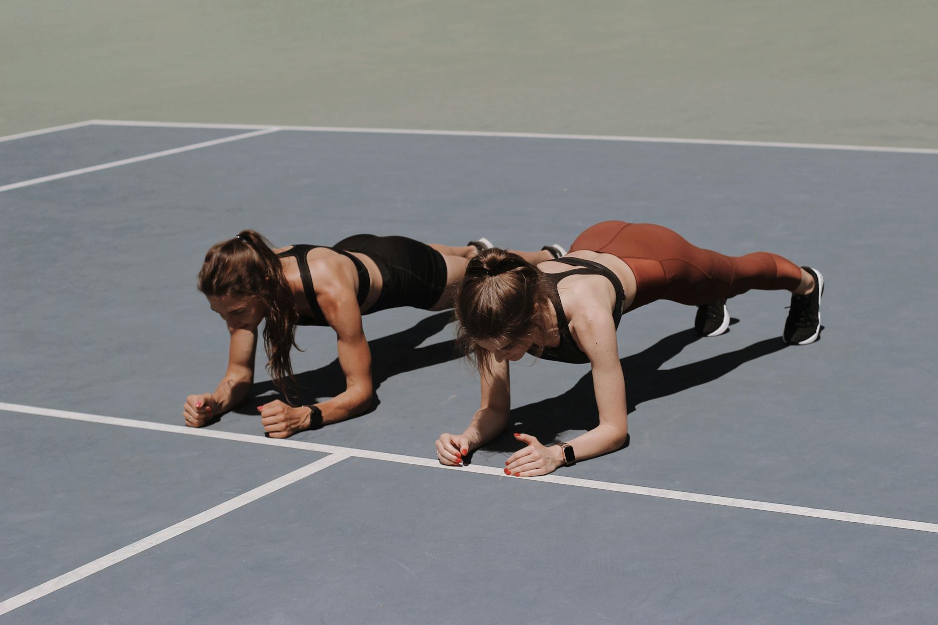 Two women in activewear doing plank exercises on an outdoor sports court. - 4 day workout routine
