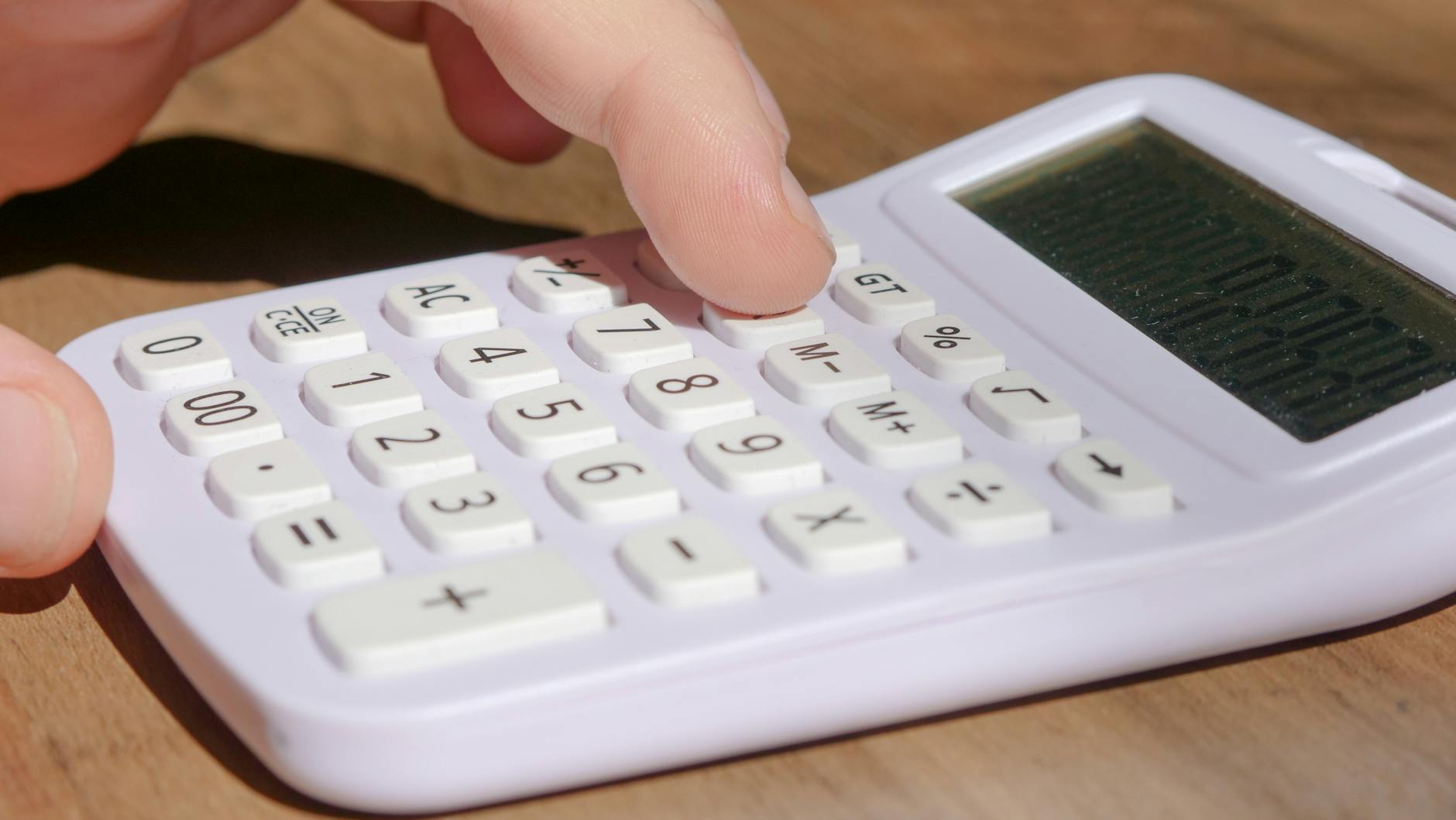 A hand pressing keys on a white calculator placed on a wooden desk in bright daylight. - calorie deficit calculator
