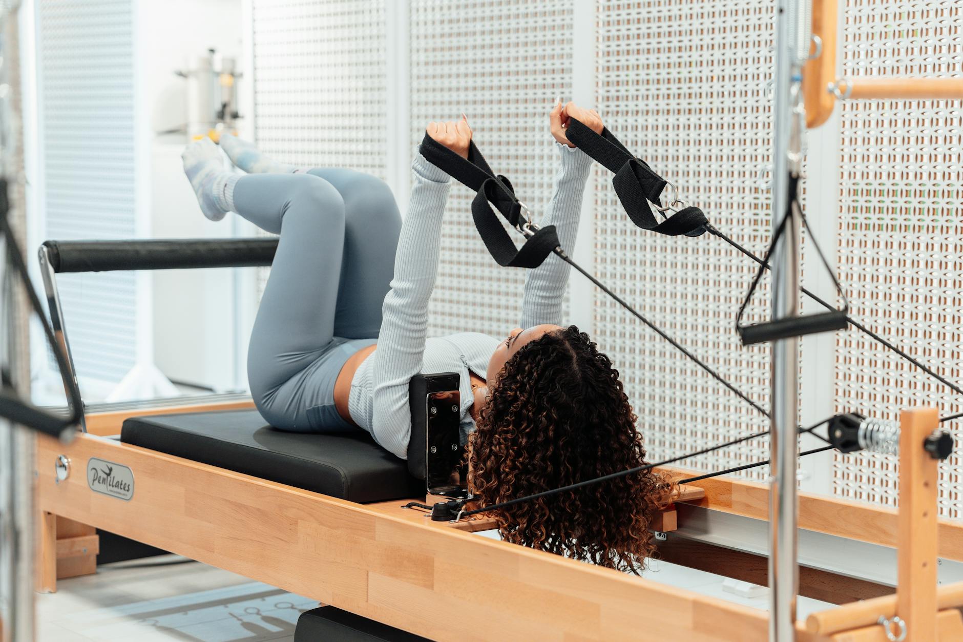 A woman wearing a gray tracksuit exercises on a Pilates reformer in a modern studio setting. - common pilates exercises