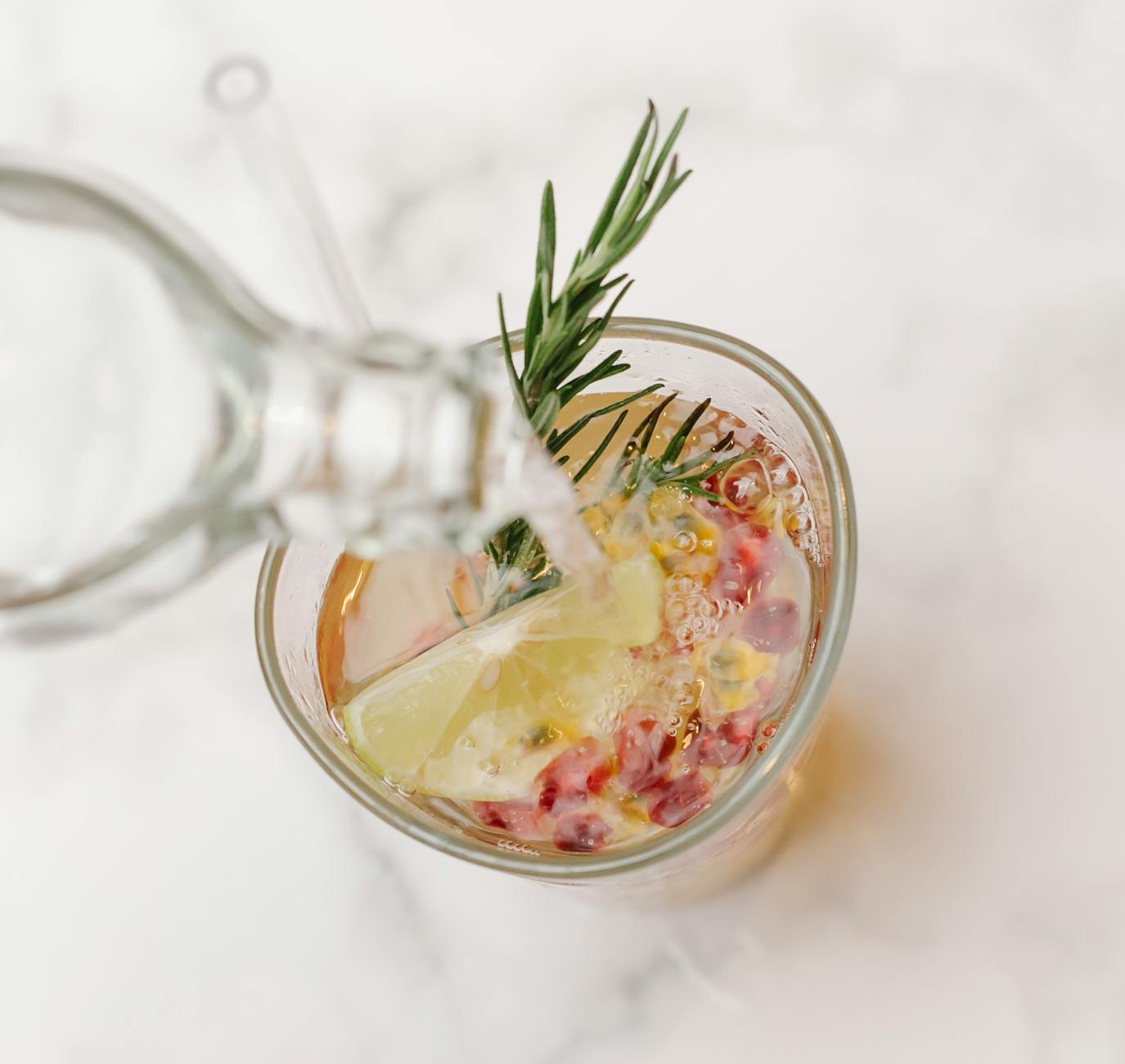 From above of glass bottle pouring water into glass with sprig of rosemary and slice of lemon placed on marble surface - detox water recipes