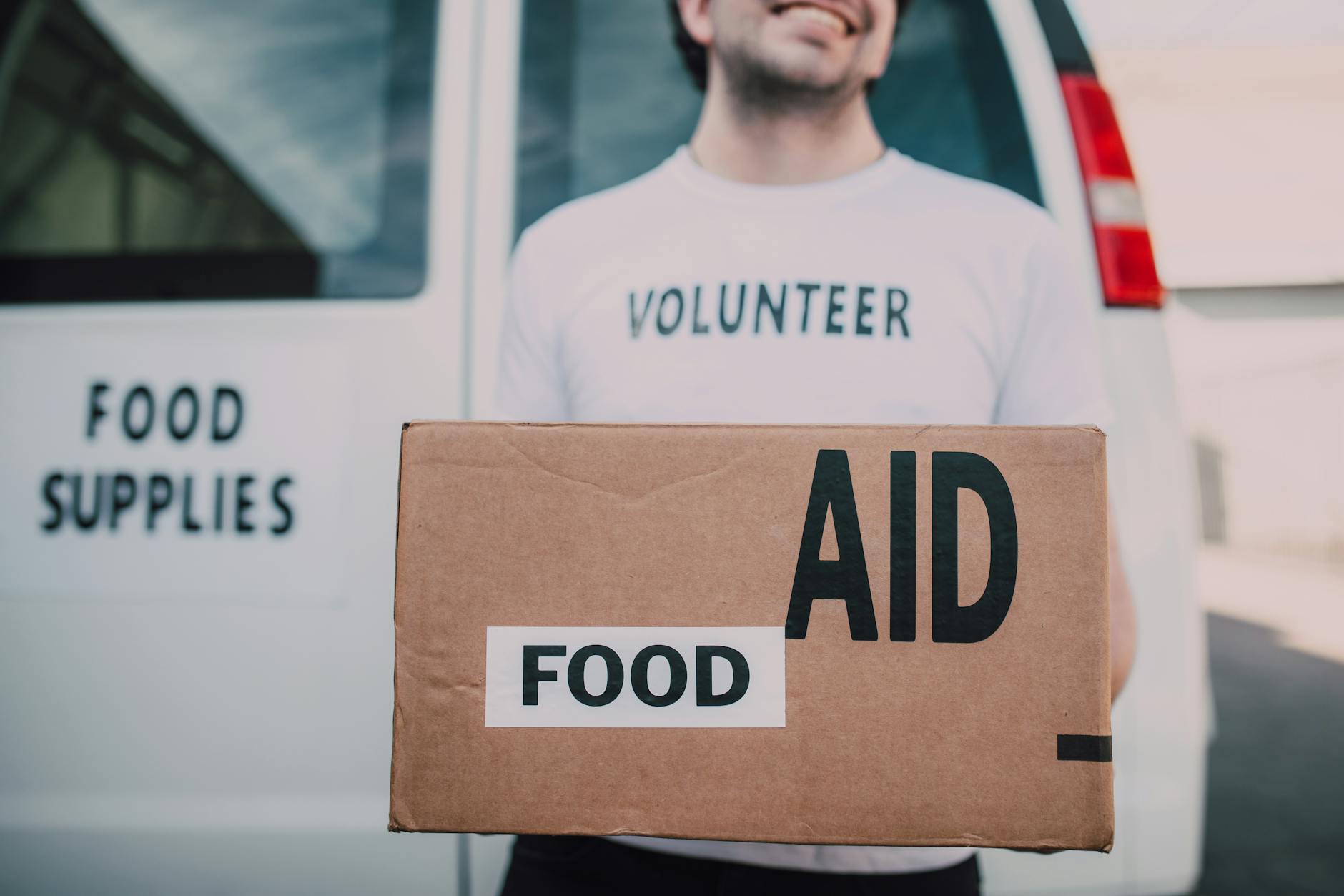 Smiling volunteer holding a box labeled 'Food Aid' outside a van with 'Food Supplies' sign. - foods that cause inflammation