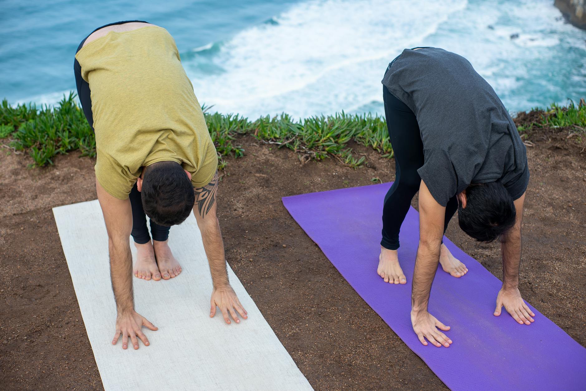 Two men practicing yoga outdoors by the sea in Portugal, performing forward bends on yoga mats. - forward head posture exercises
