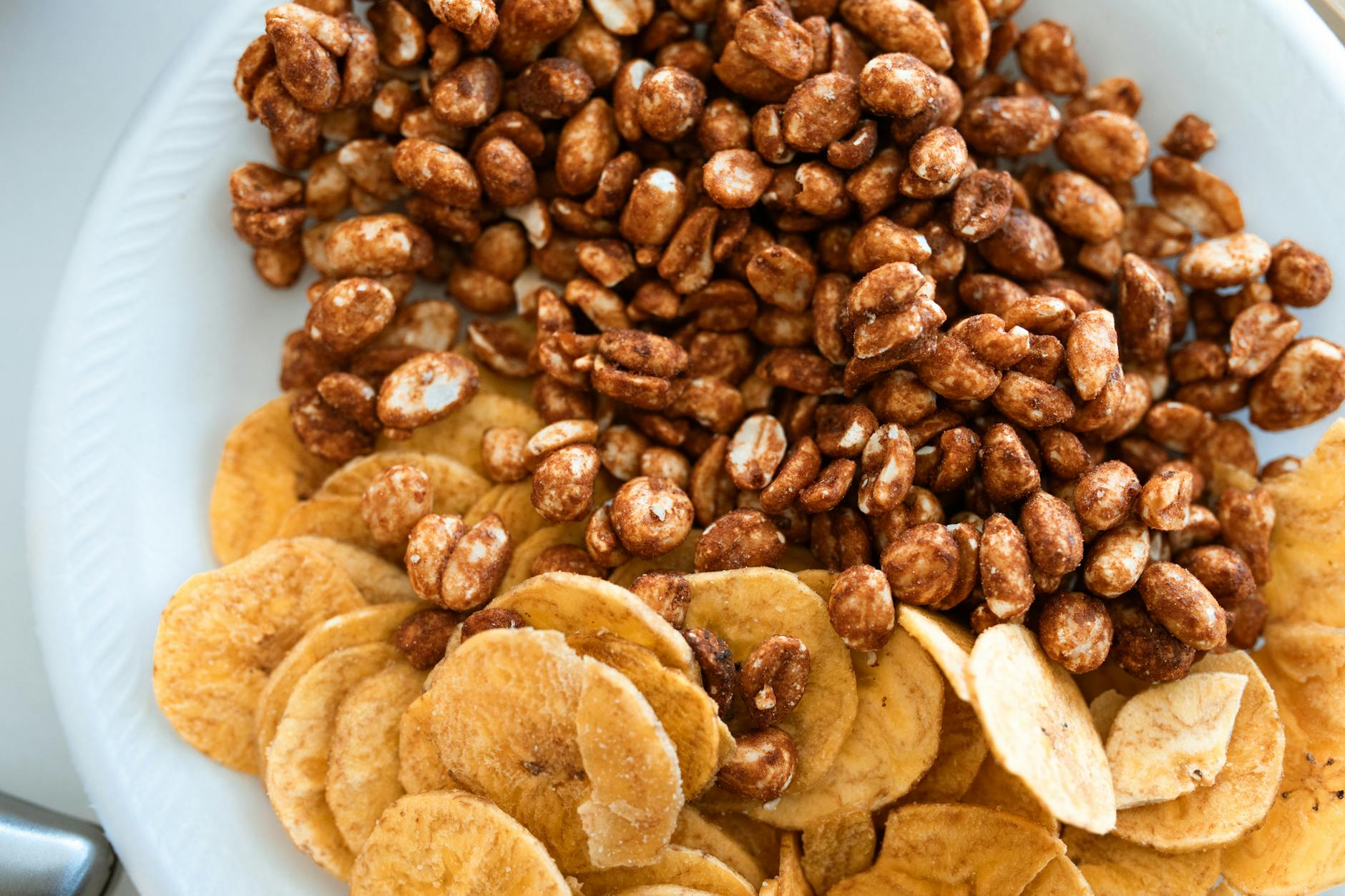 Close-up image of crispy banana chips and sweet honey roasted peanuts on a ceramic plate. - healthy snack ideas