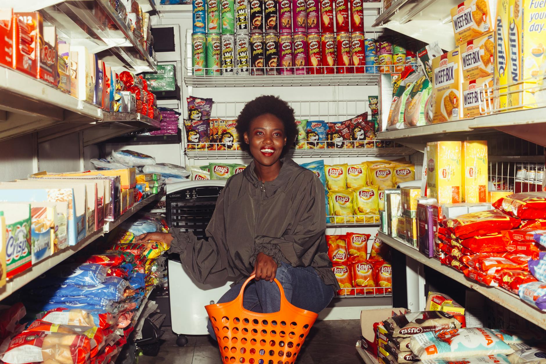 Full length of cheerful ethnic woman in casual clothes with shopping bag squatting among store shelves choosing food for buying - healthy snacks to buy