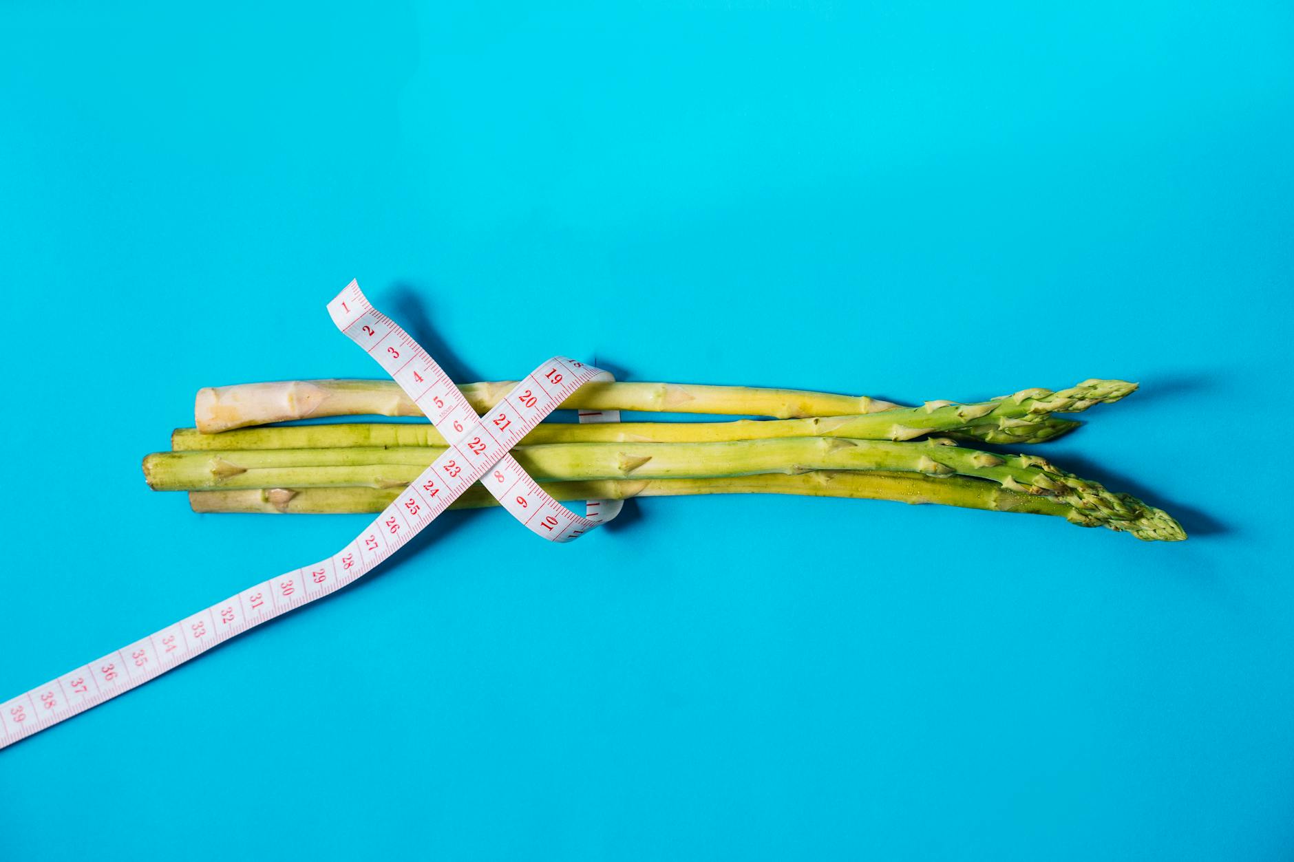 Fresh asparagus wrapped with measuring tape on a vibrant blue background. - low calorie deficit
