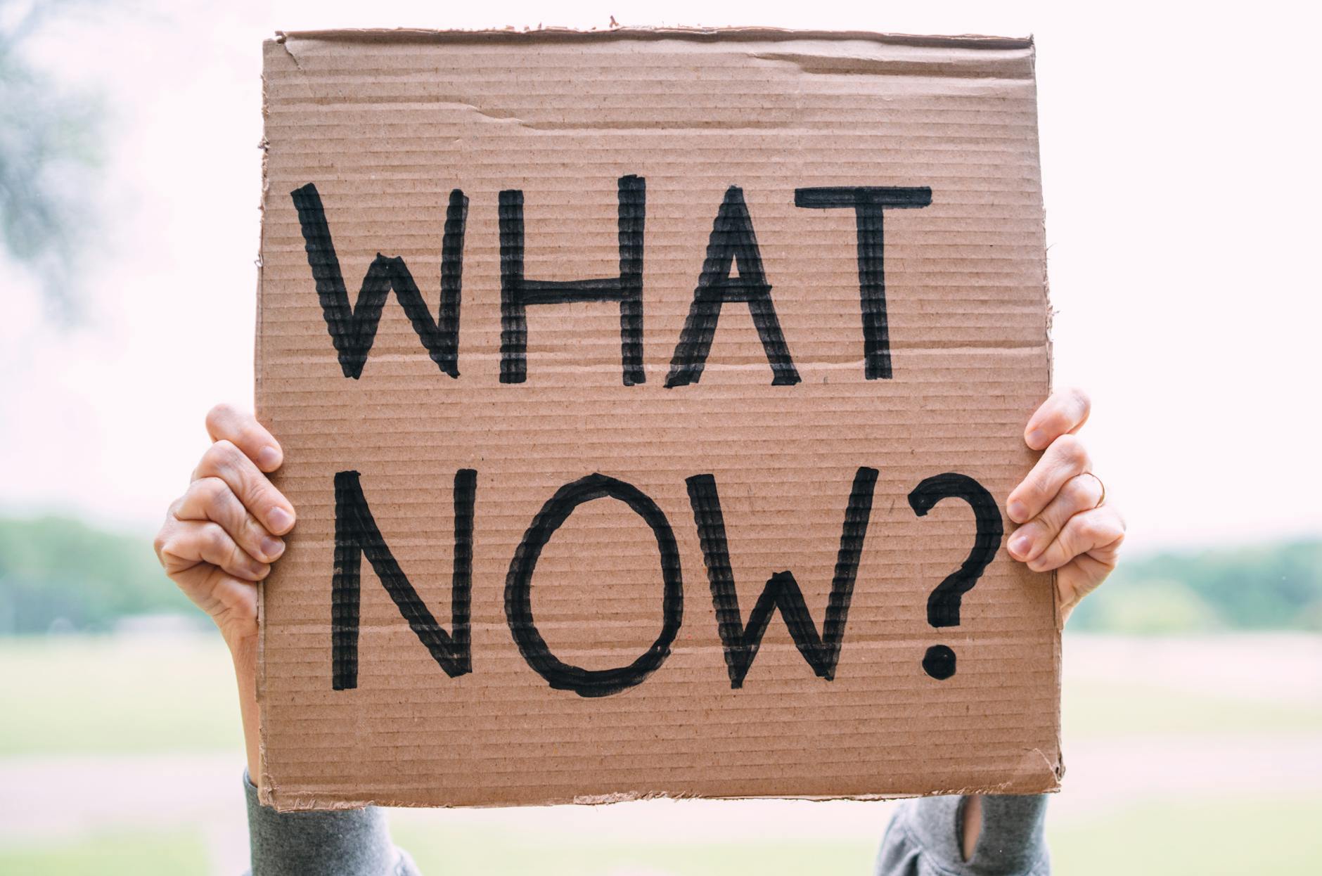 Cardboard sign reading 'What Now?' held outdoors, conveying uncertainty or protest. - what is meditation