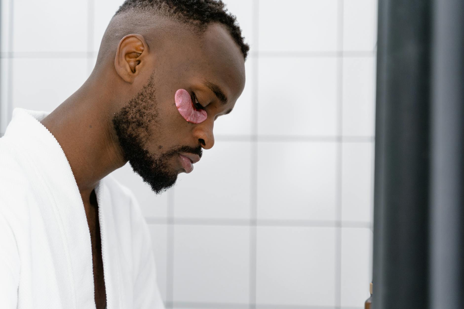 African American man in white robe applying under eye mask for skincare routine. - men's natural skincare