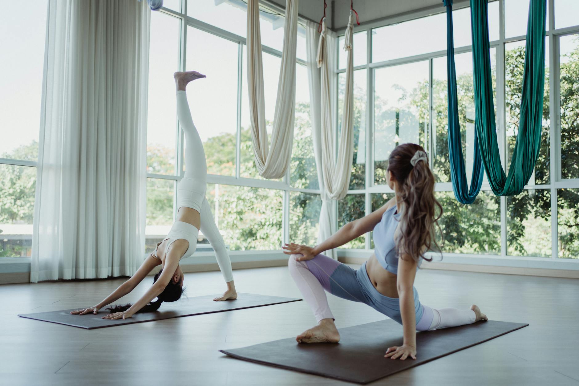 Two women practicing yoga poses in a bright, airy studio with aerial silks and large windows. - shoulder flexibility exercises