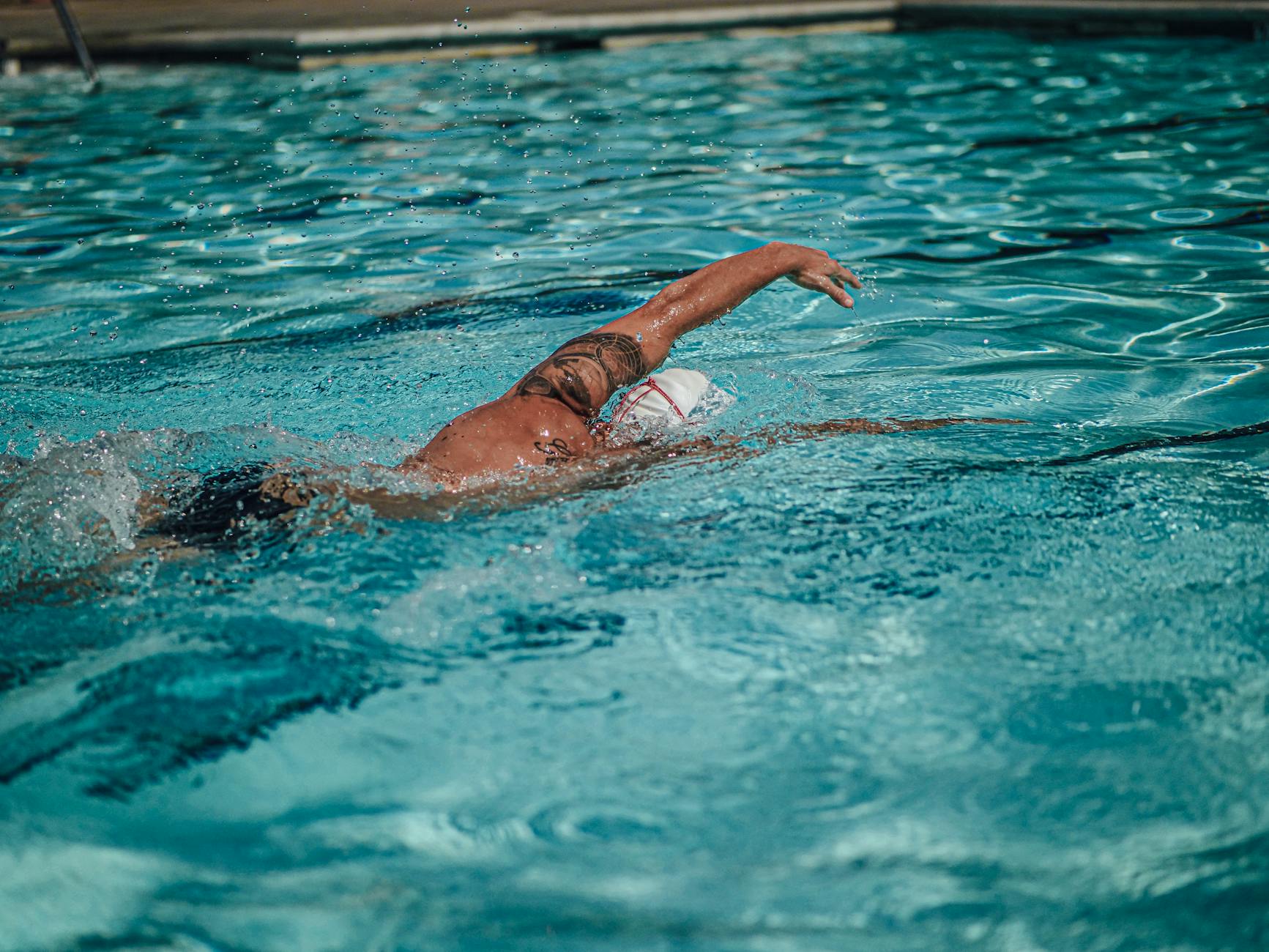 A tattooed swimmer practicing freestyle stroke in a sunlit swimming pool. - swimming benefits for men