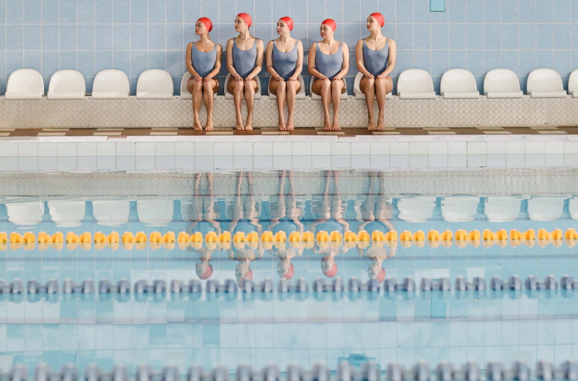 Five women in blue swimsuits sit poolside wearing red swim caps, ready for water sports training. - swimming benefits women