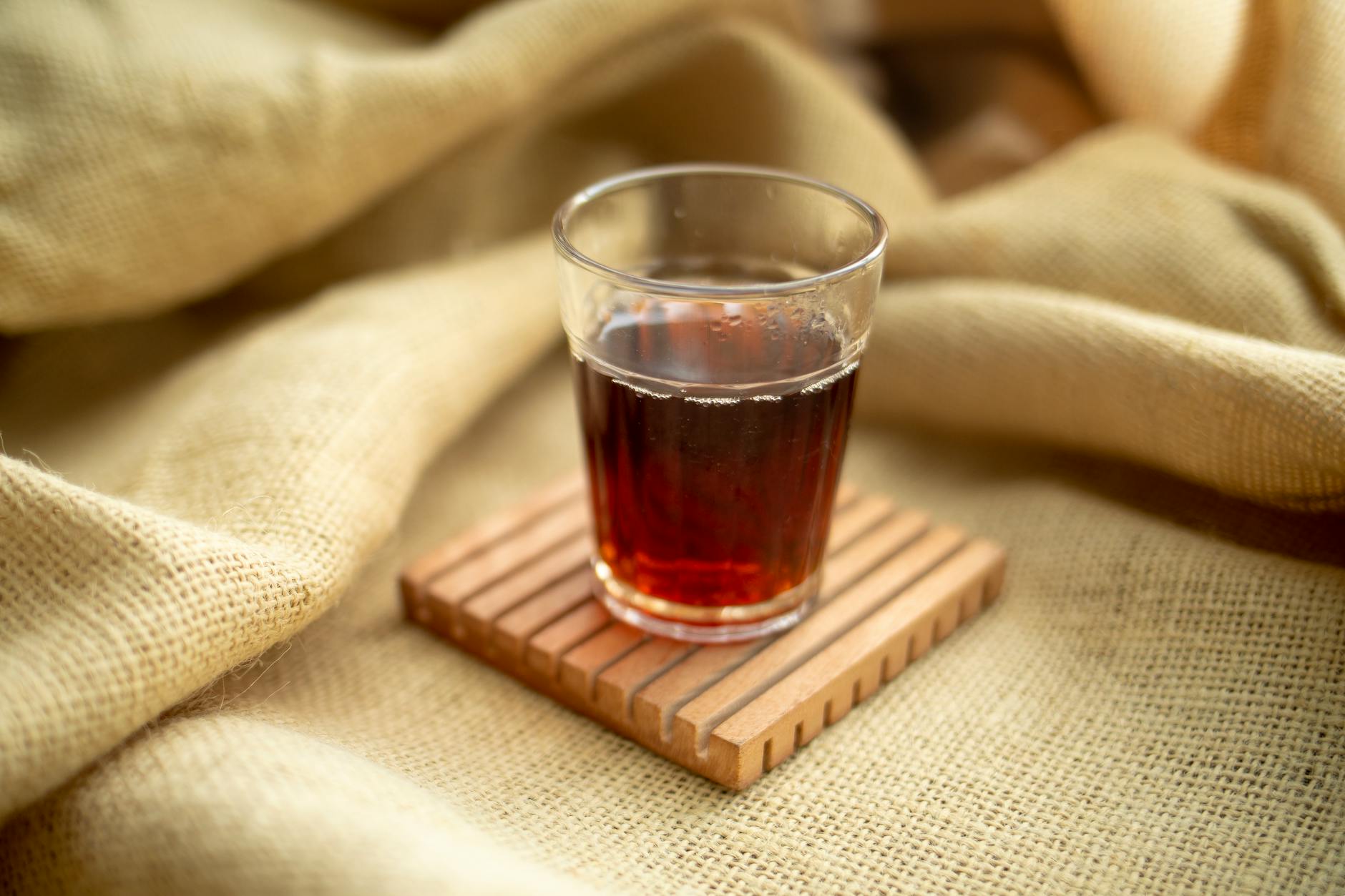 A clear glass filled with warm tea resting on a wooden coaster and burlap fabric. - yoga classes near me