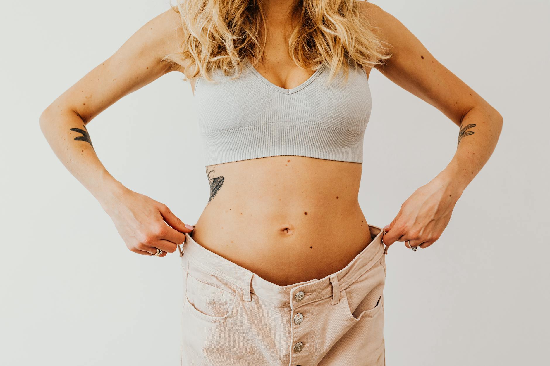 A woman displaying weight loss by wearing oversized pants and crop top, showing tattoos. - belly fat workout