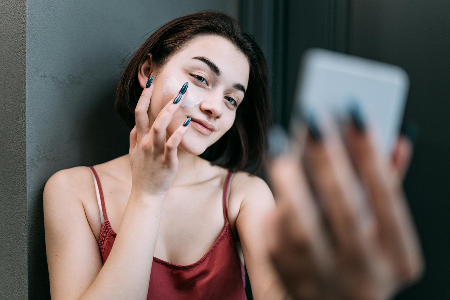 Young Caucasian woman applying moisturizer while smiling in front of a mirror indoors. - best natural moisturizer