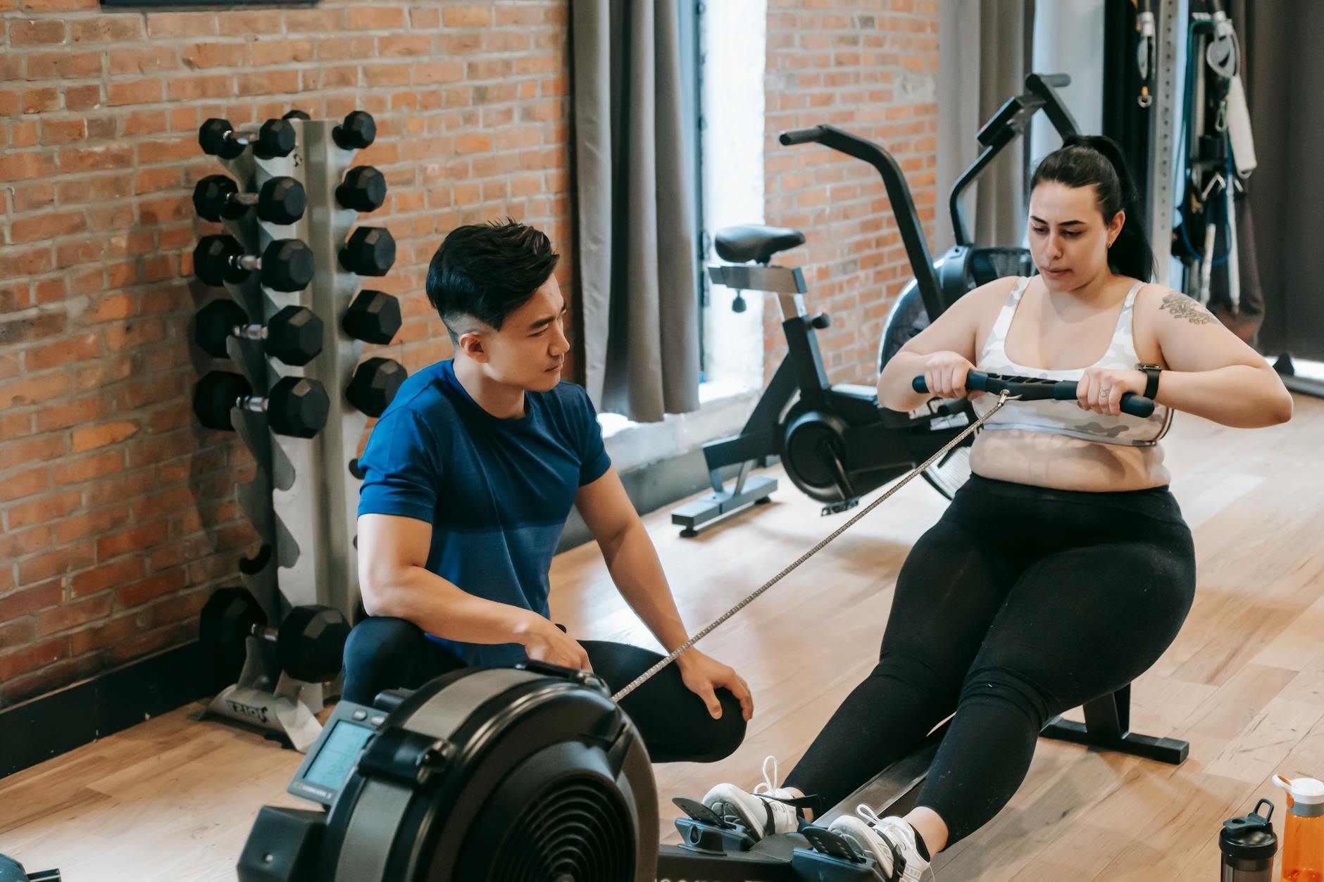 From above of professional ethnic constructor helping young plus size woman working out with rowing machine in sport club - cardio fat burning