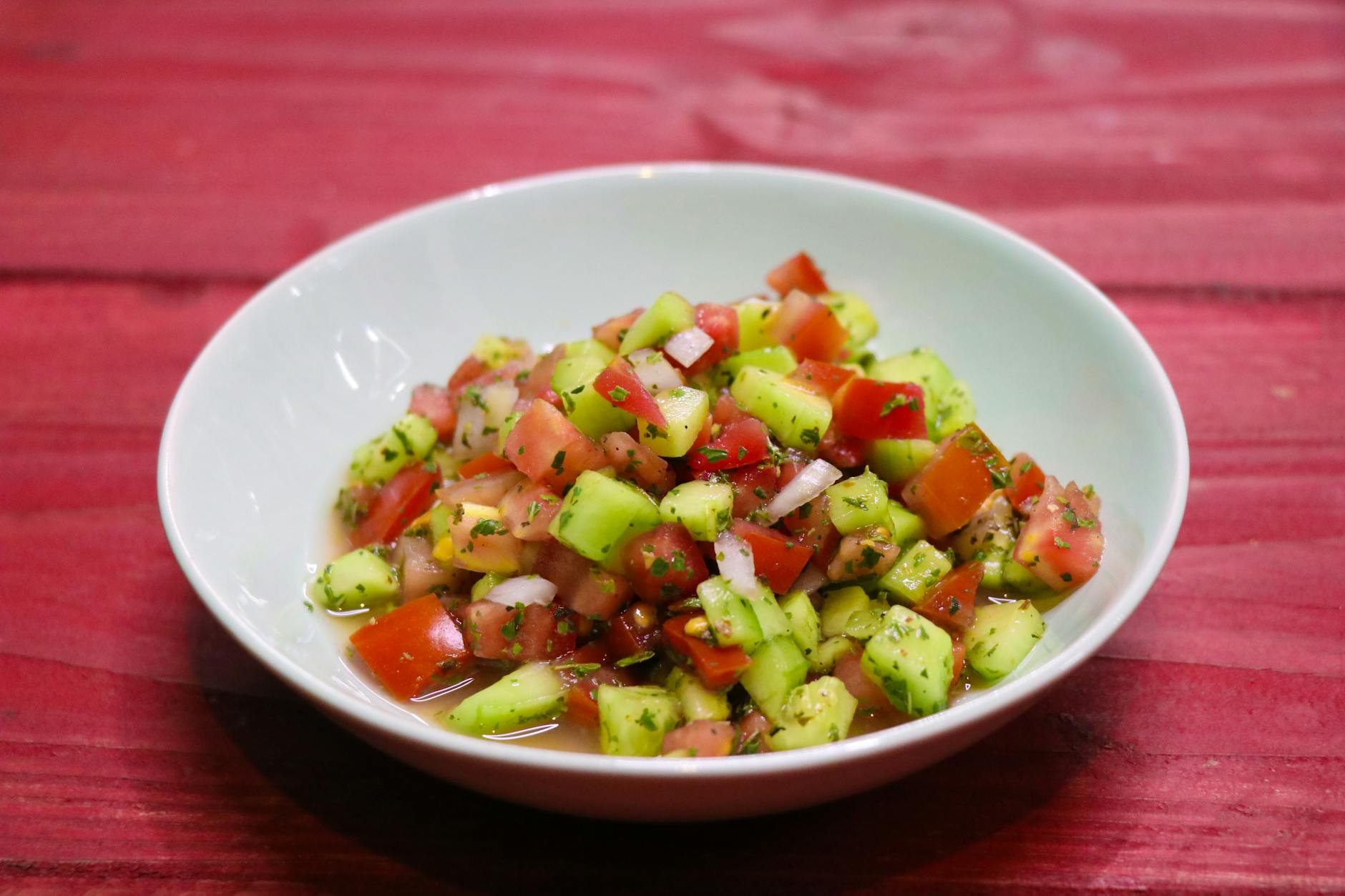 A vibrant cucumber and tomato salad in a white bowl, set on a red wooden table. - easy healthy meals