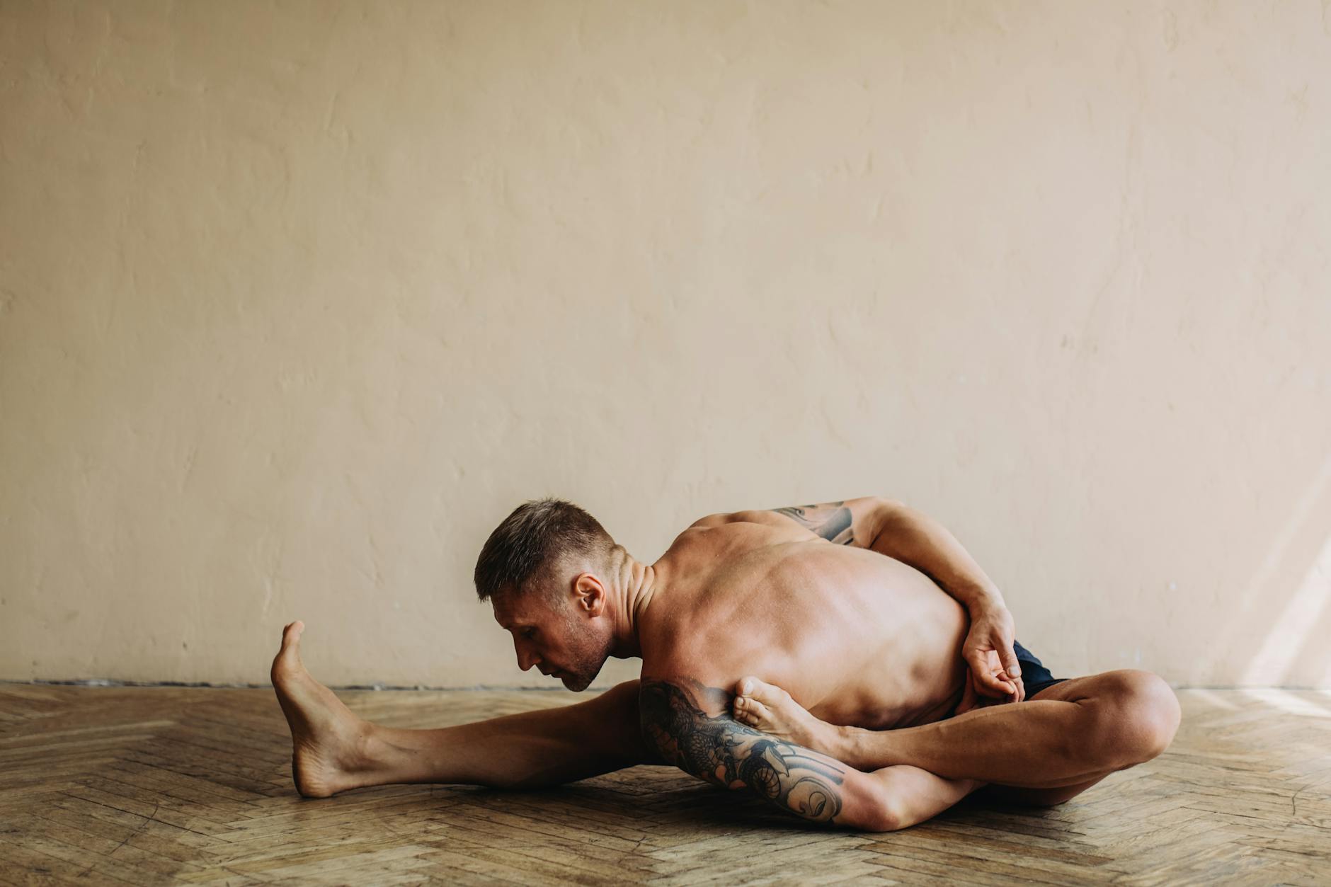 Tattooed man practicing a challenging yoga pose indoors, showcasing flexibility and strength. - full body stretch