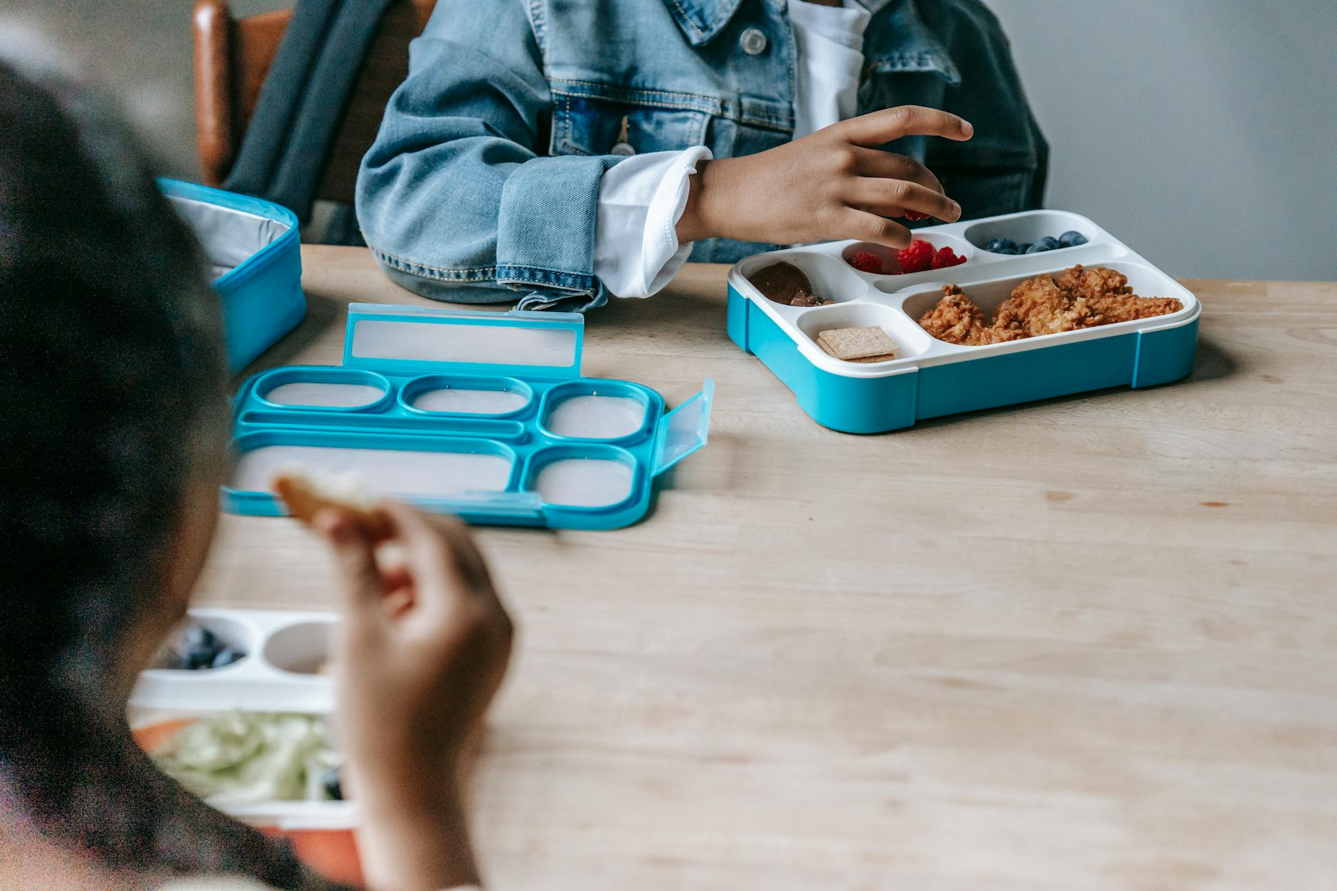 Crop faceless classmates sitting at table with lunch boxes with various food and eating during break - healthy kids lunches