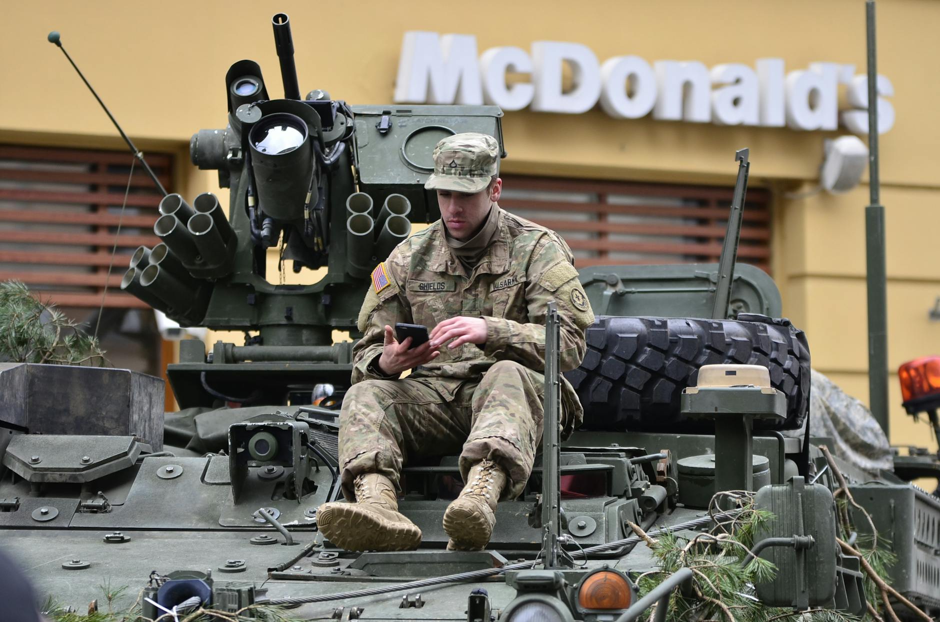 A soldier in camouflage resting on an armored vehicle, using a smartphone outside a McDonald's. - healthy mcdonalds breakfast