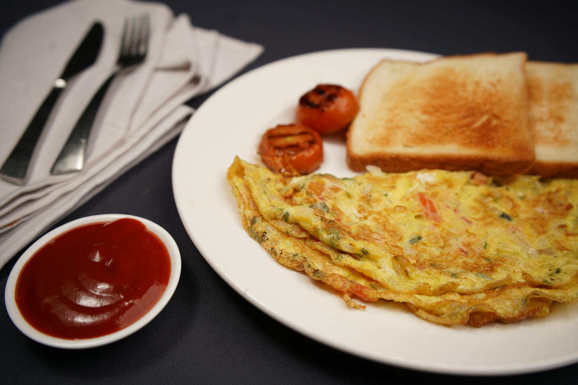 A hearty breakfast featuring omelette, toast, grilled tomatoes, and ketchup on a black background. - high protein breakfast ideas