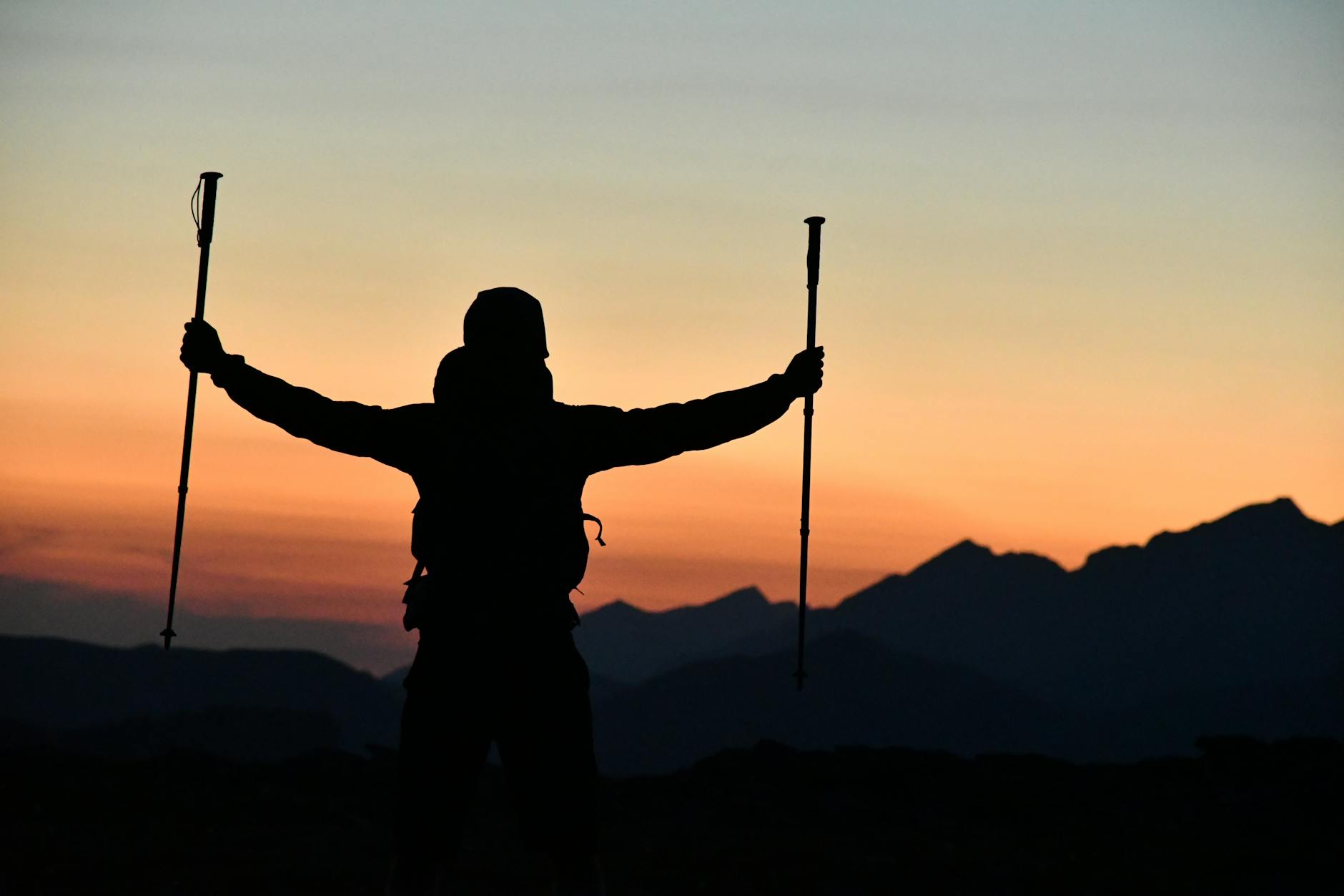 A silhouetted hiker triumphantly raises trekking poles against a breathtaking sunset mountain backdrop. - hiking calorie burn