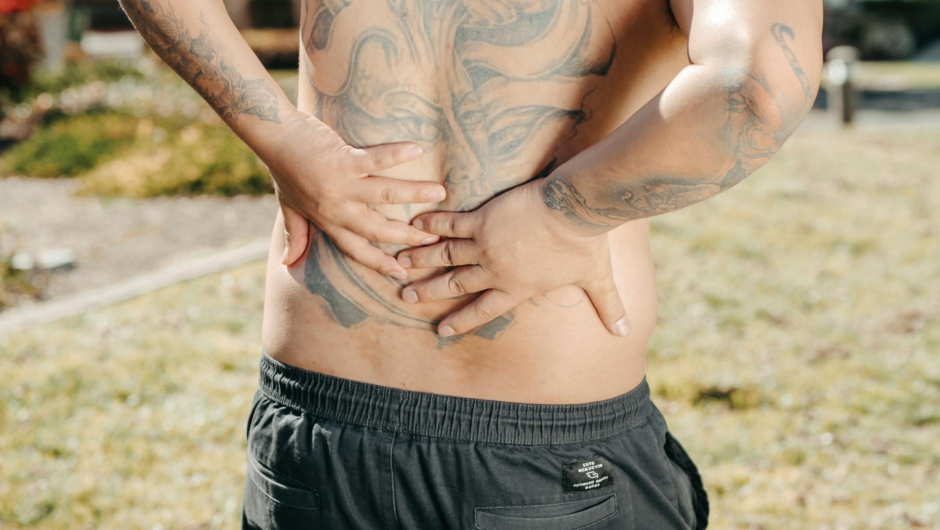 A man with a large tattoo on his back holds his lower back outdoors, showing discomfort. - lower body exercises