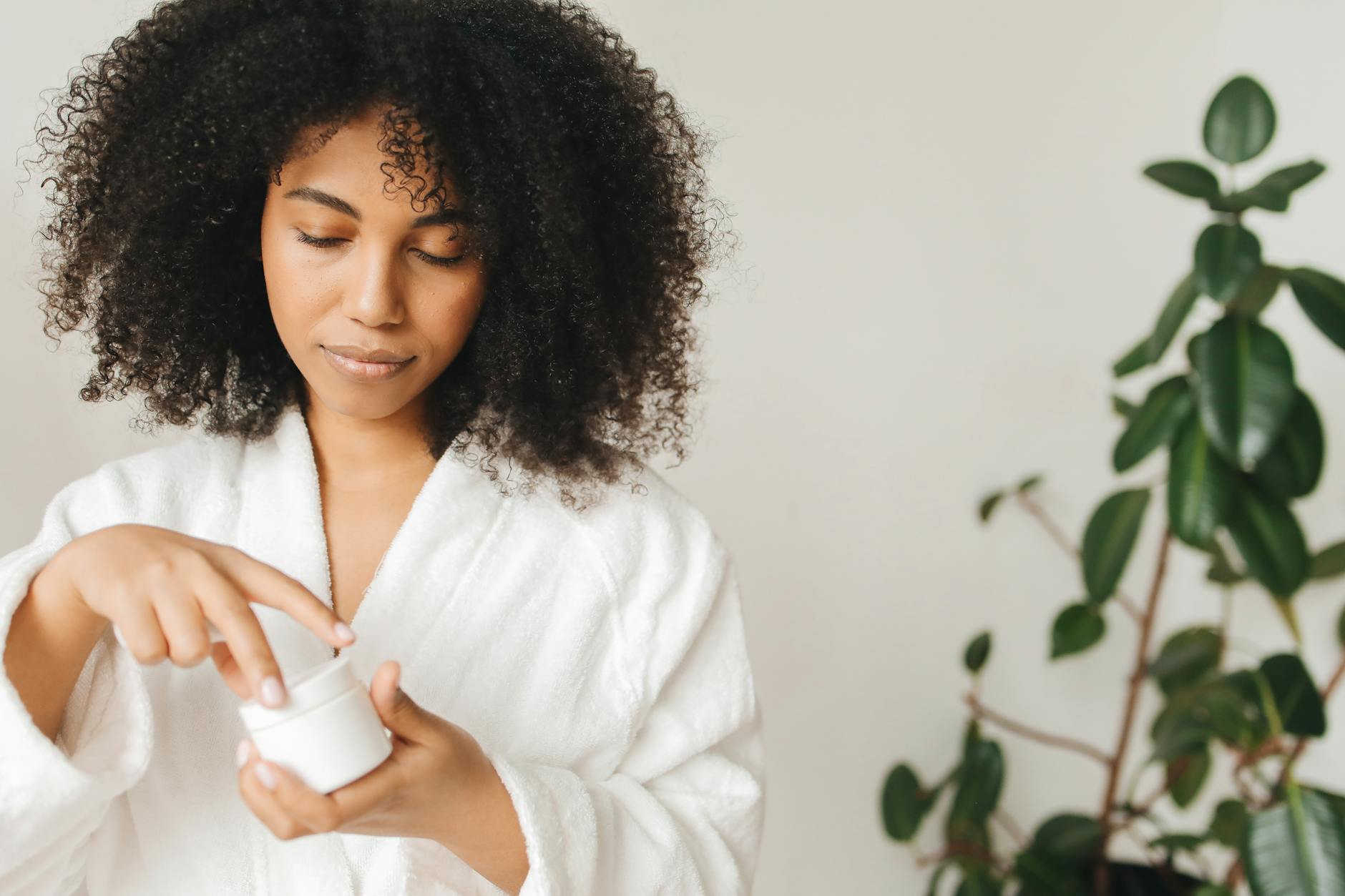 A black woman applies skincare cream indoors, wearing a white robe beside a green plant. - natural skincare routine steps