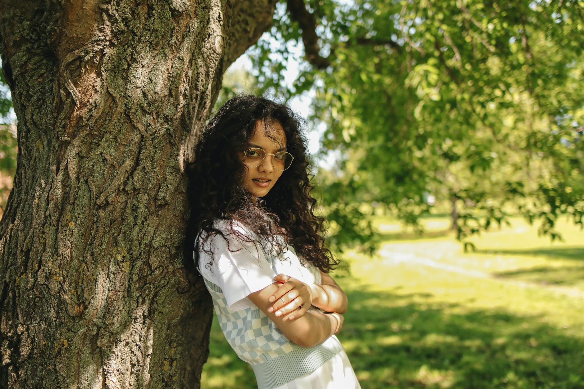 Young woman with eyeglasses posing stylishly near a tree in a sunlit park. - skinny recipes